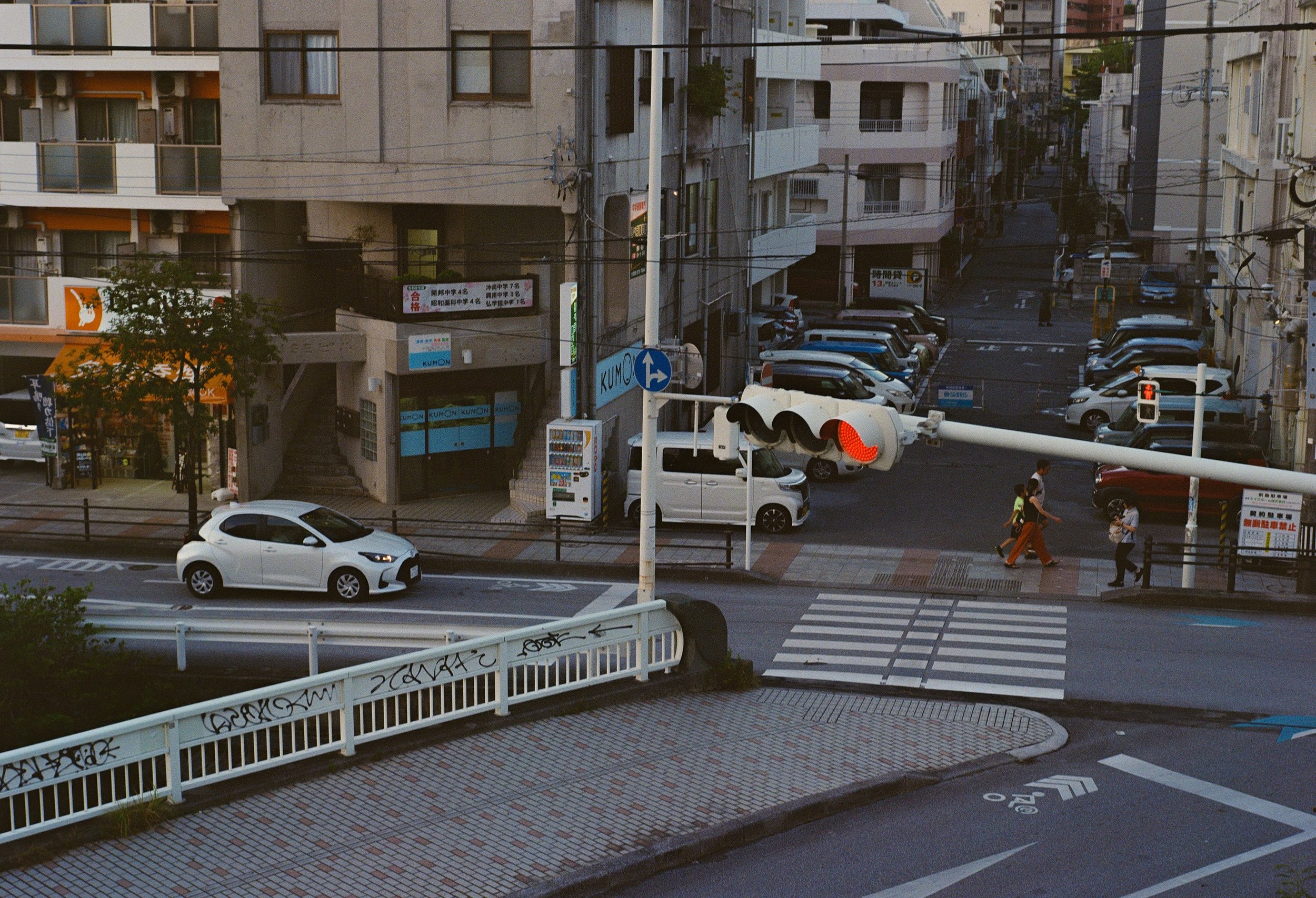 An urban street scene with cars, pedestrians, traffic lights, and buildings, including a parking lot and signs.