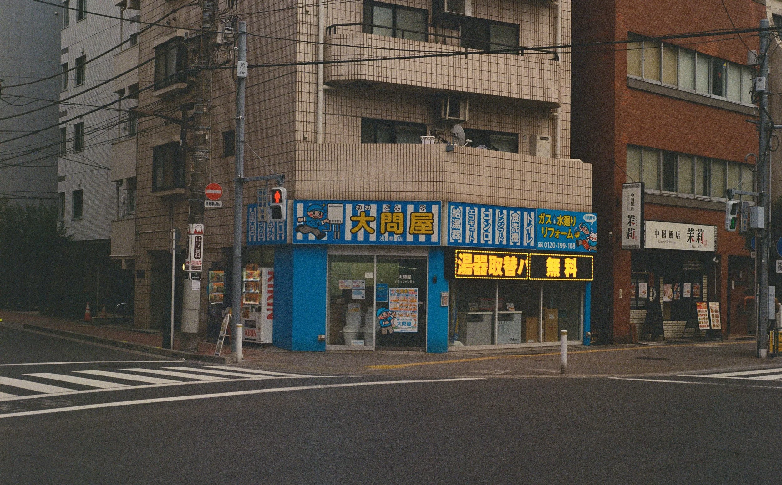 A small storefront on a city street with blue and yellow signage in Japanese, vending machines outside, and a digital sign with Japanese characters. There are crosswalk lines on the street in front.