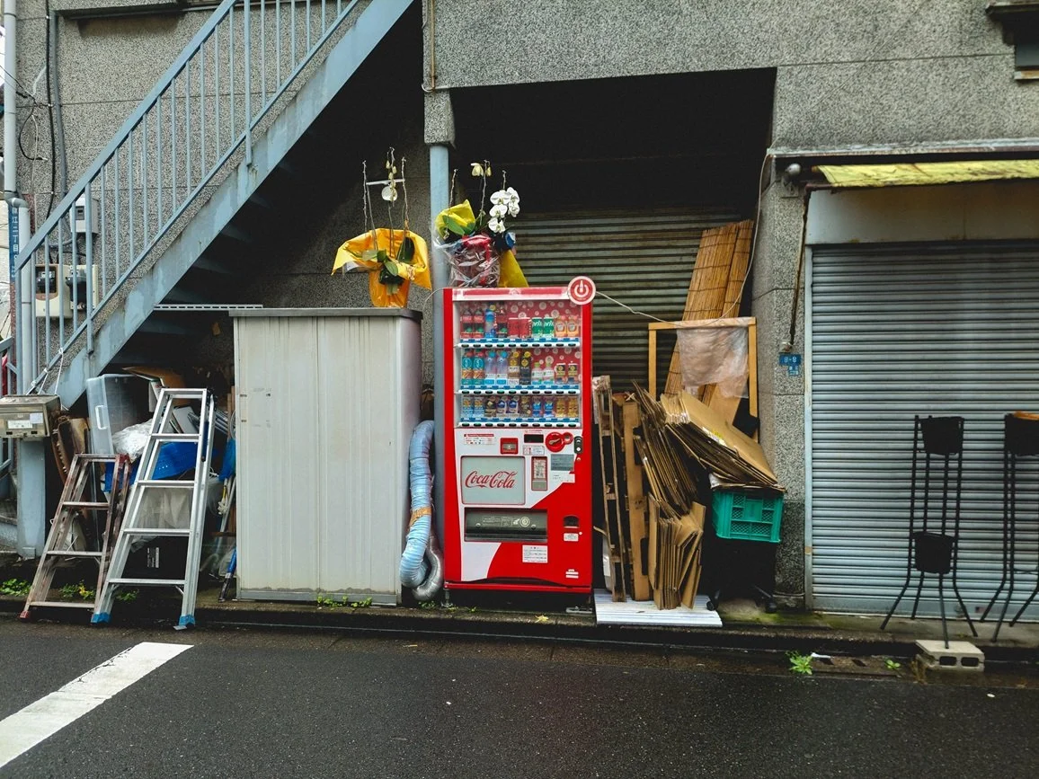 A Coca-Cola vending machine standing outside near a gray metal garage door, with cardboard boxes, metal racks, and flower arrangements on top of the vending machine, next to a set of exterior stairs.