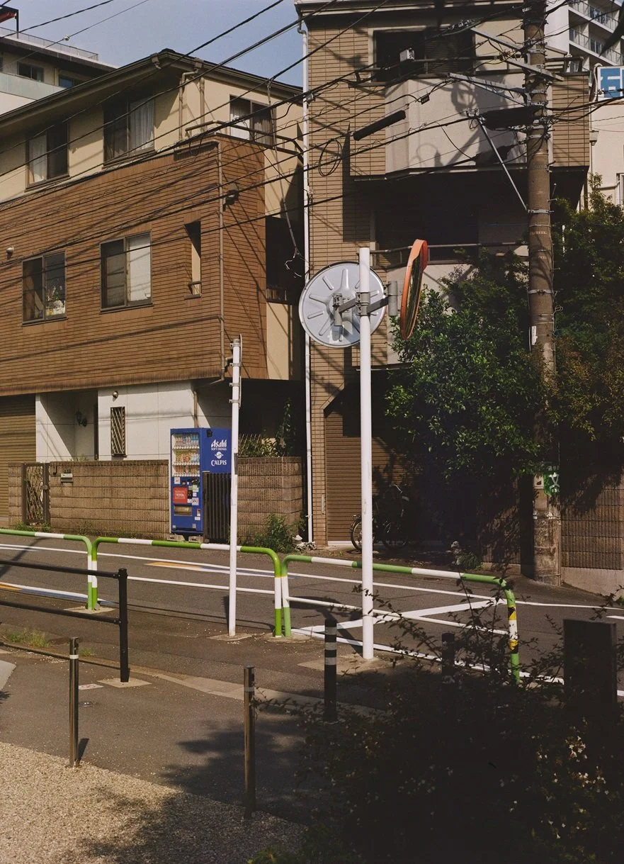 A street scene featuring a brown multi-story building, a vending machine on the sidewalk, a large clock, and overhead power lines, with some trees and a residential area.