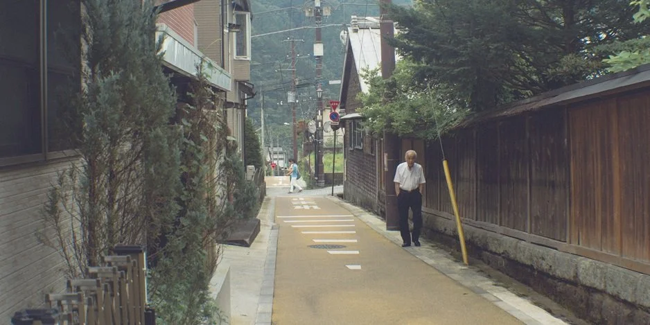 A narrow sidewalk in a quiet residential area with an older man walking along it, surrounded by wooden fences and houses, with trees and parked bicycles visible.