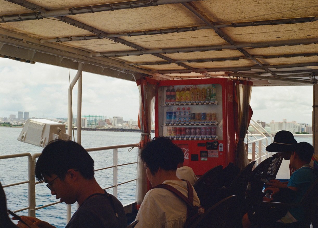 People sitting on a boat with a vending machine on board, overlooking a river and city skyline in the background.