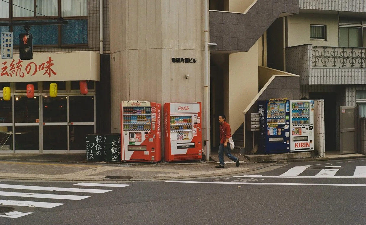 A city street scene with four vending machines lined up on the sidewalk near a building corner. A man is walking past the vending machines, wearing a red jacket and carrying a bag. There is a crosswalk in the foreground, and the building has Japanese