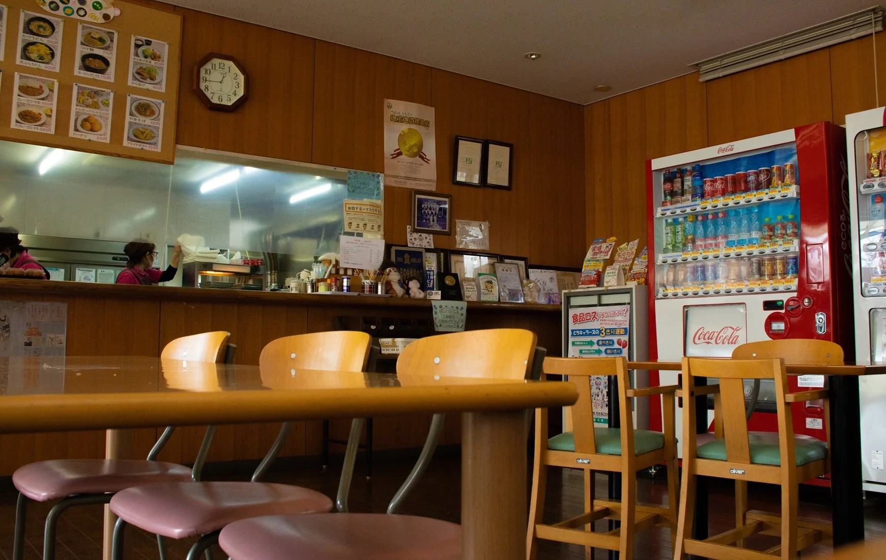 Interior of a small restaurant with wooden tables and chairs, a vending machine, a counter with kitchen staff, and framed awards on the wall.