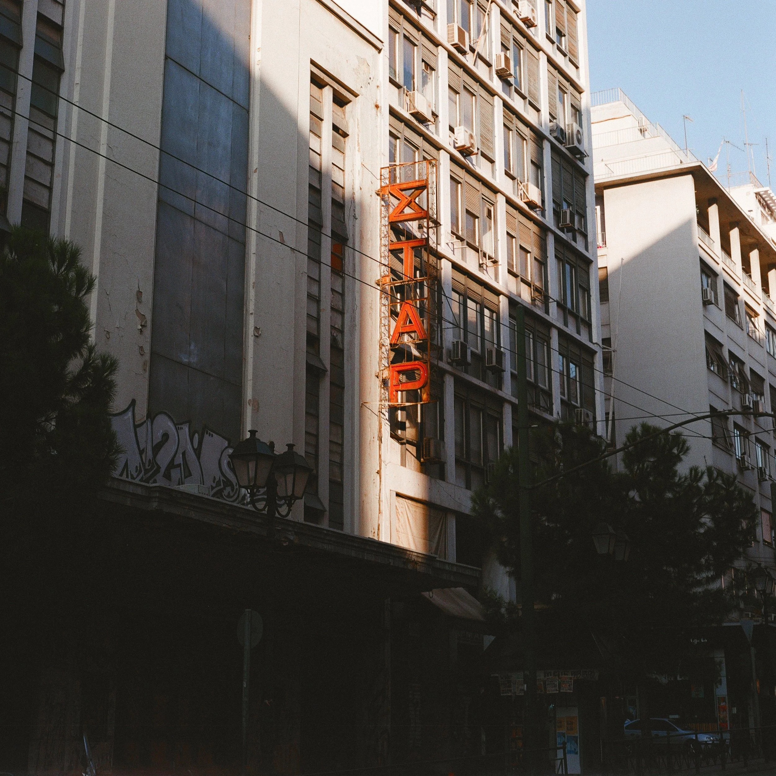 An old vertical neon sign reading 'ZITTA RAD' attached to the exterior of a tall, worn building with multiple air conditioning units in windows, city street scene with trees and street lamps