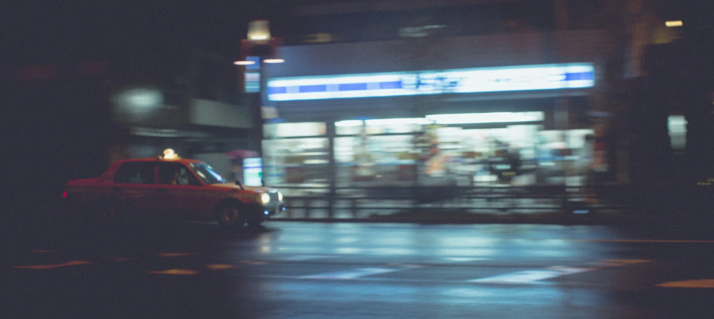 A red taxi driving past a brightly lit storefront at night with a blurred background.