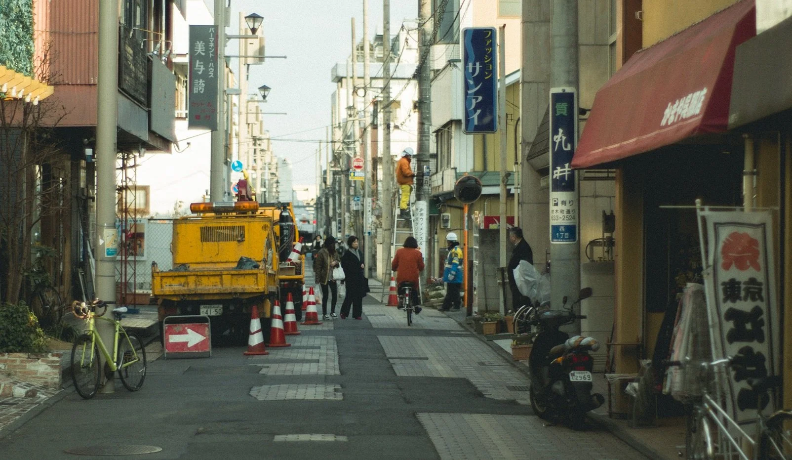 A city street with people, bicycles, a construction vehicle, and shop signs.