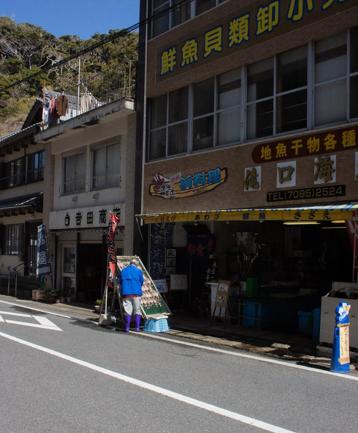 A street scene in Japan with a person selling seafood in front of a building with Japanese signs. The person is wearing a blue jacket and purple boots.