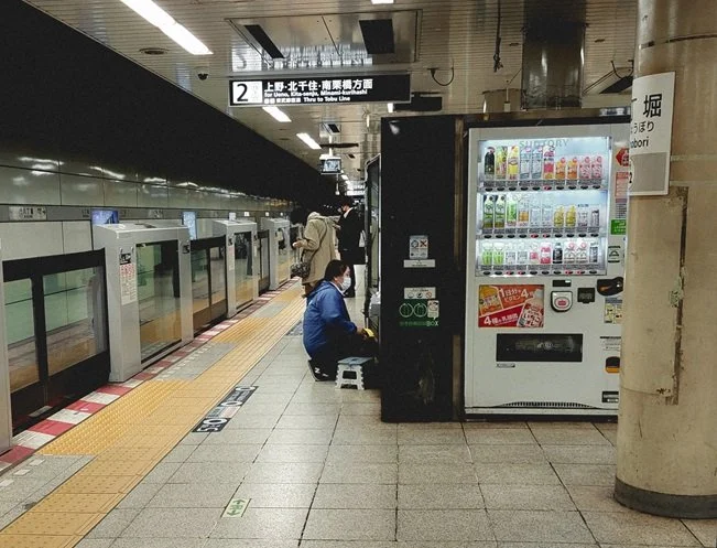People waiting on a subway platform with a vending machine beside a safety barrier.