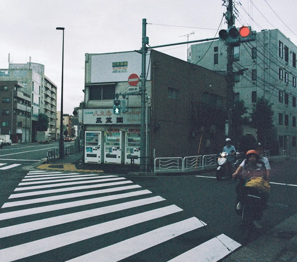 City street corner with crosswalk, vending machines, traffic light, and motorcyclists with helmets riding on the street.