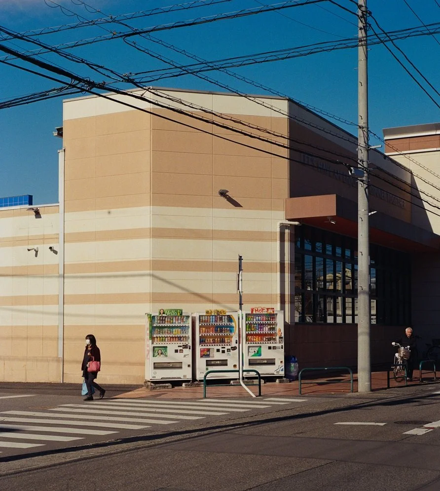 Street scene with two people, vending machines, utility pole, and power lines outside a beige building under a clear blue sky.