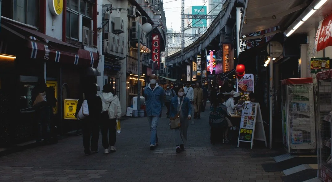 Street scene in a busy shopping district with pedestrians walking, illuminated signs in Japanese, storefronts, and outdoor vendors.