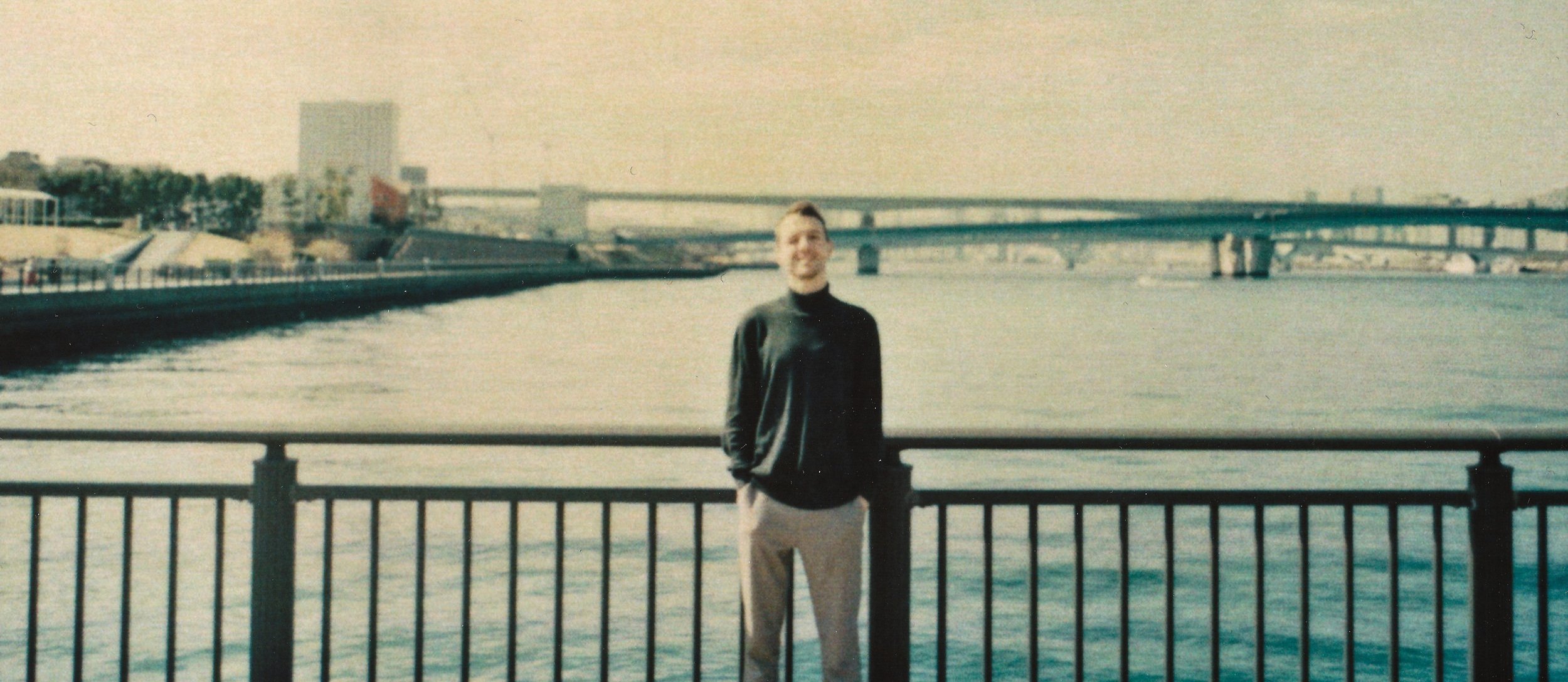 A young man standing by a waterfront railing with a cityscape and bridge in the background. this is me. edo. furikakedo.