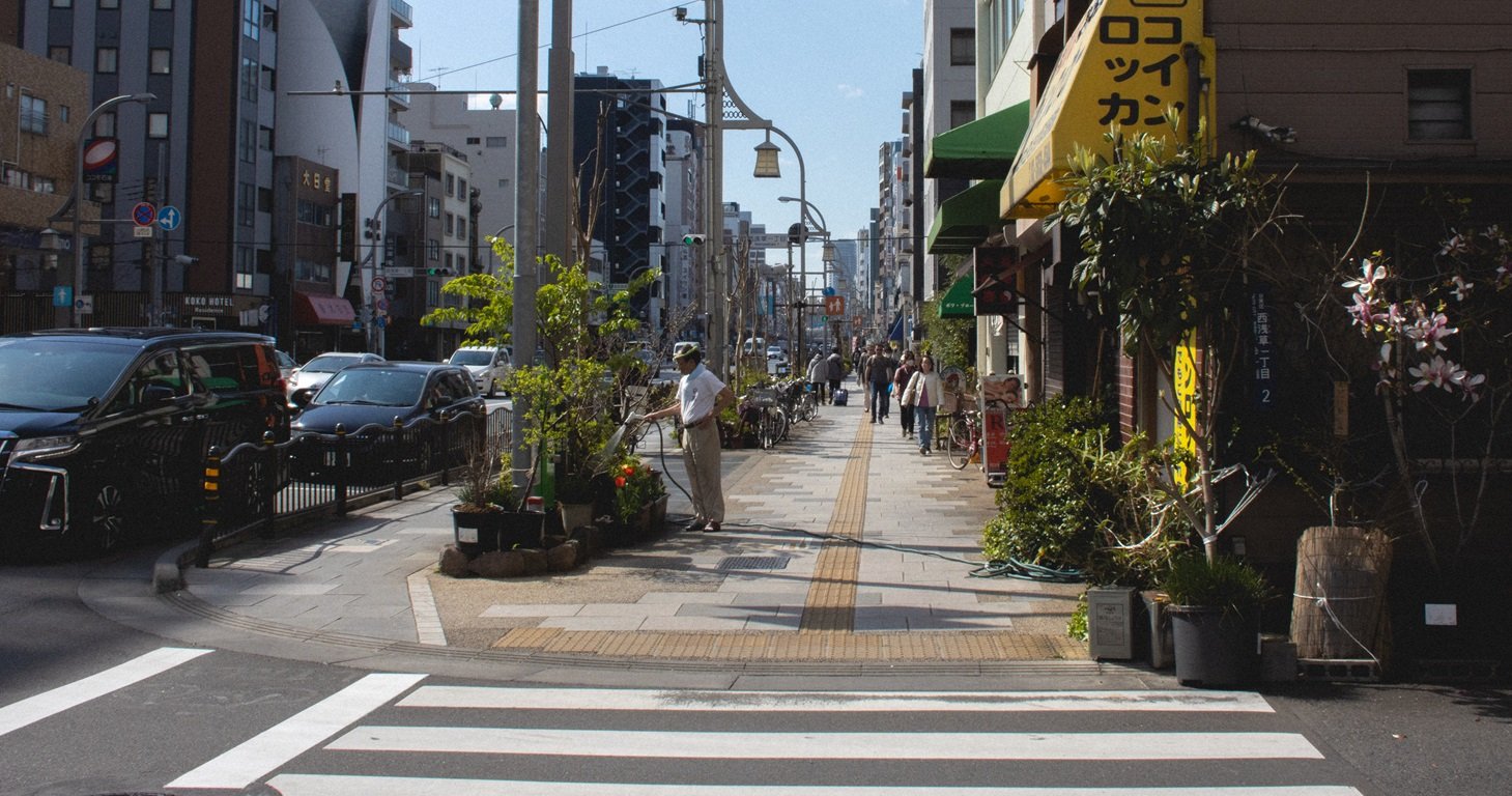 City sidewalk with pedestrians, shops, and parked bicycles, with cars on the street and buildings in the background.