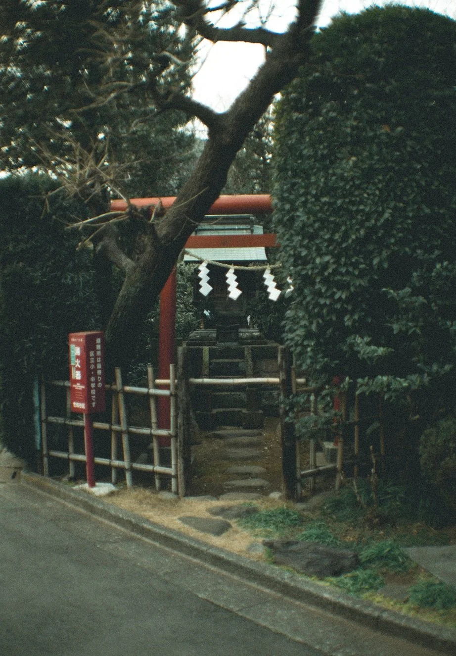 A small Japanese shrine entrance with a red torii gate, surrounded by trees and shrubbery, with steps leading up to the shrine.
