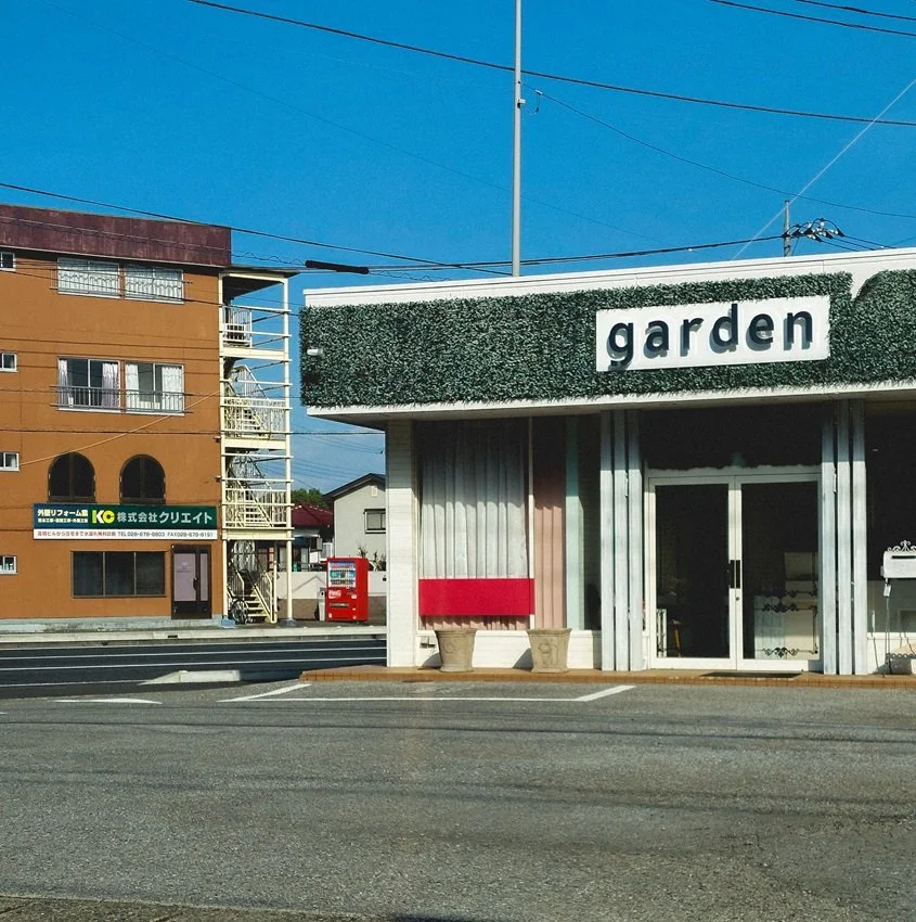 A building with a sign reading 'garden' and a green hedge above the entrance. The building has a glass door and two beige planters outside. The street in front is empty, and there are power lines overhead.