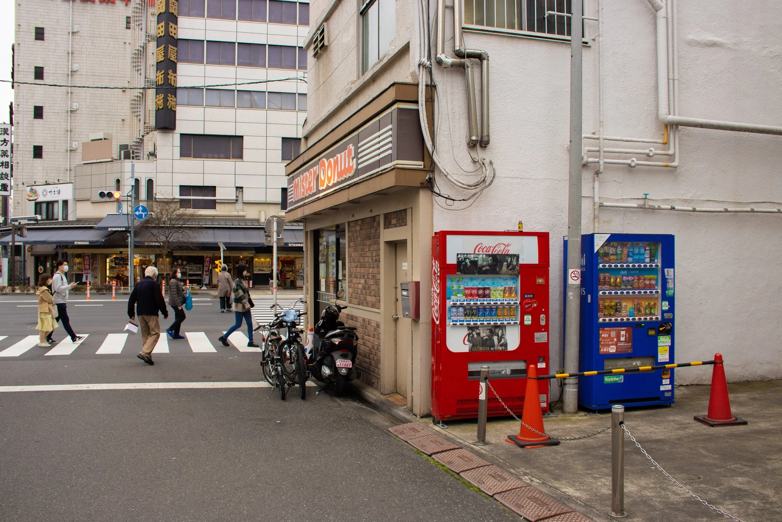 City street corner with vending machines and pedestrians walking across crosswalks. Two vending machines are located against a building on the right, one red Coca-Cola machine and one blue snack machine. Several bicycles and a scooter are parked near