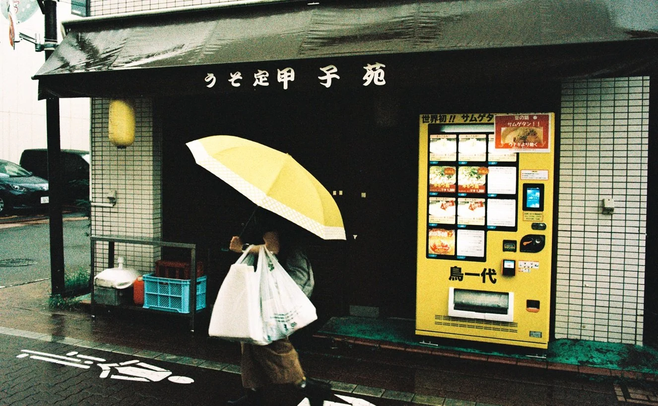 A woman walking past a Japanese vending machine holding shopping bags and an umbrella on a rainy day.