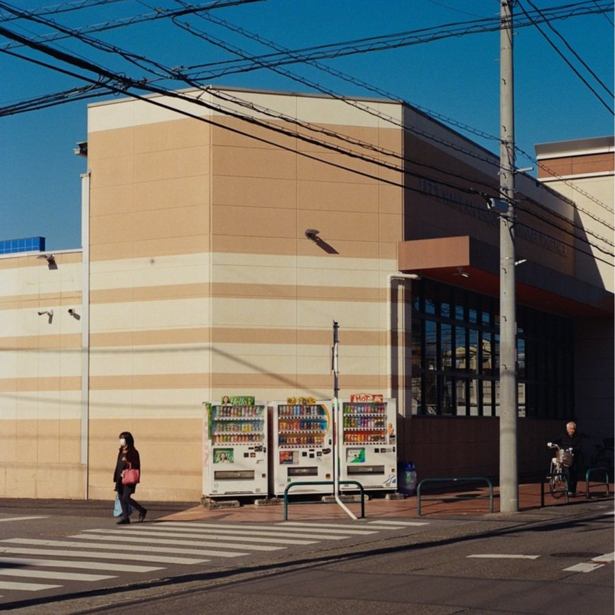 Urban scene with a large beige building, power lines overhead, two vending machines, a woman walking with a mask carrying a pink bag, and a man sitting on a bench with a bicycle on a bright sunny day.