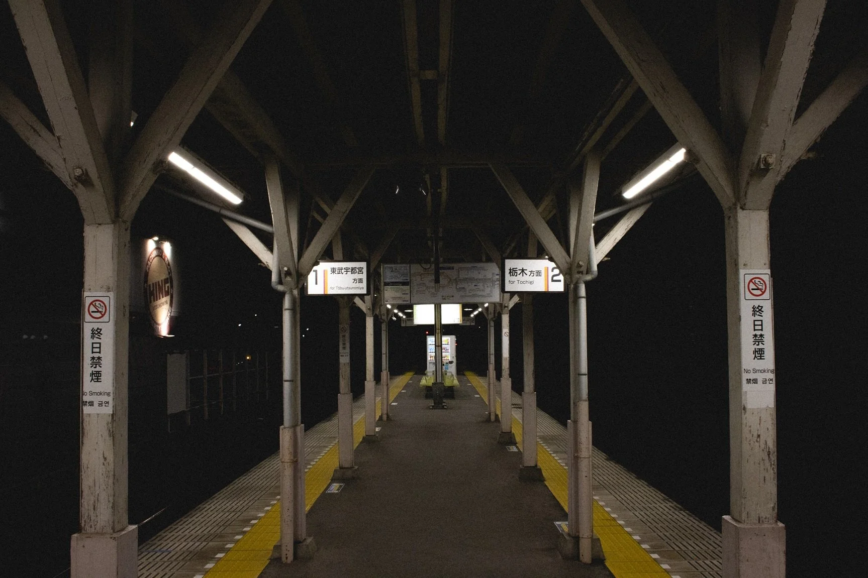 An empty train station platform at night with signs for Tobu Utsunomiya Line and Tohchi Line. No people are visible, and there are notices for no smoking. The platform has yellow tactile paths for the visually impaired.
