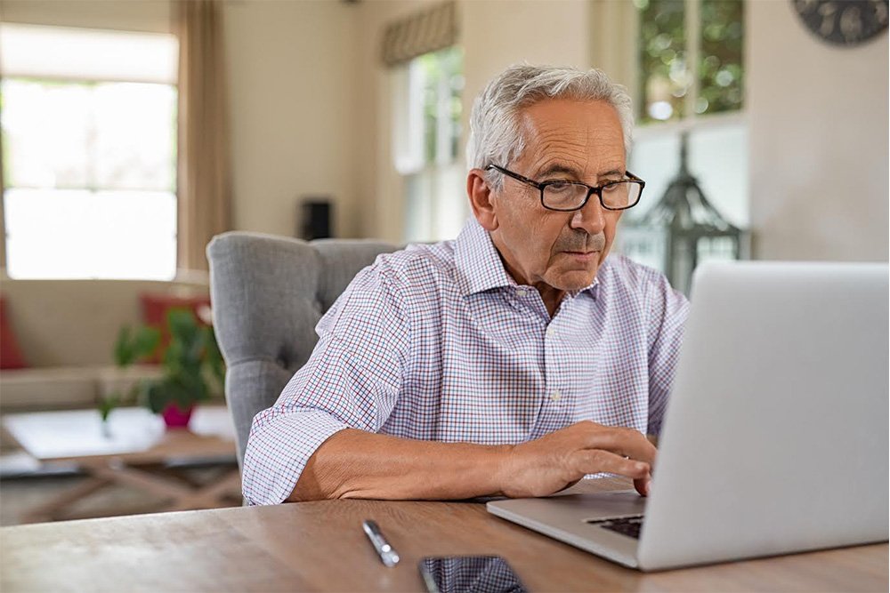 Older man with glasses working on a laptop at a desk in a bright room.