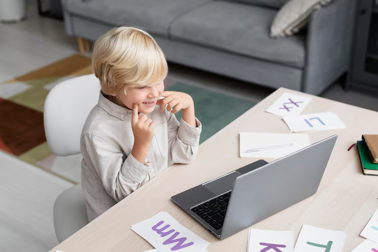 A young boy sitting at a desk, smiling and looking at a laptop with paper flashcards around him.