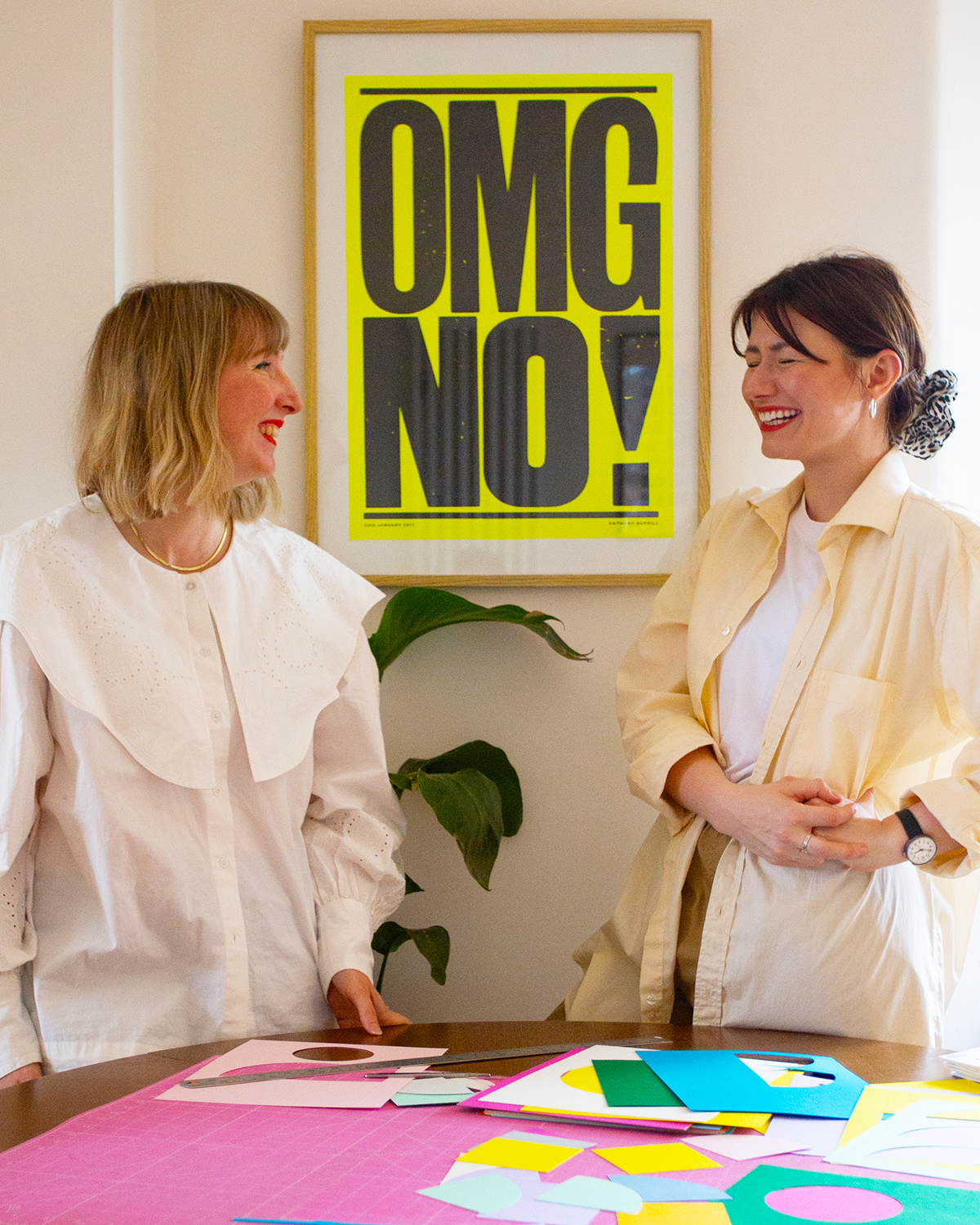 Two women with light skin are smiling and talking in a room decorated with a bright yellow sign that says 'OMG NO!'. They are standing near a table covered with colorful paper craft materials, and there's a green plant behind them.