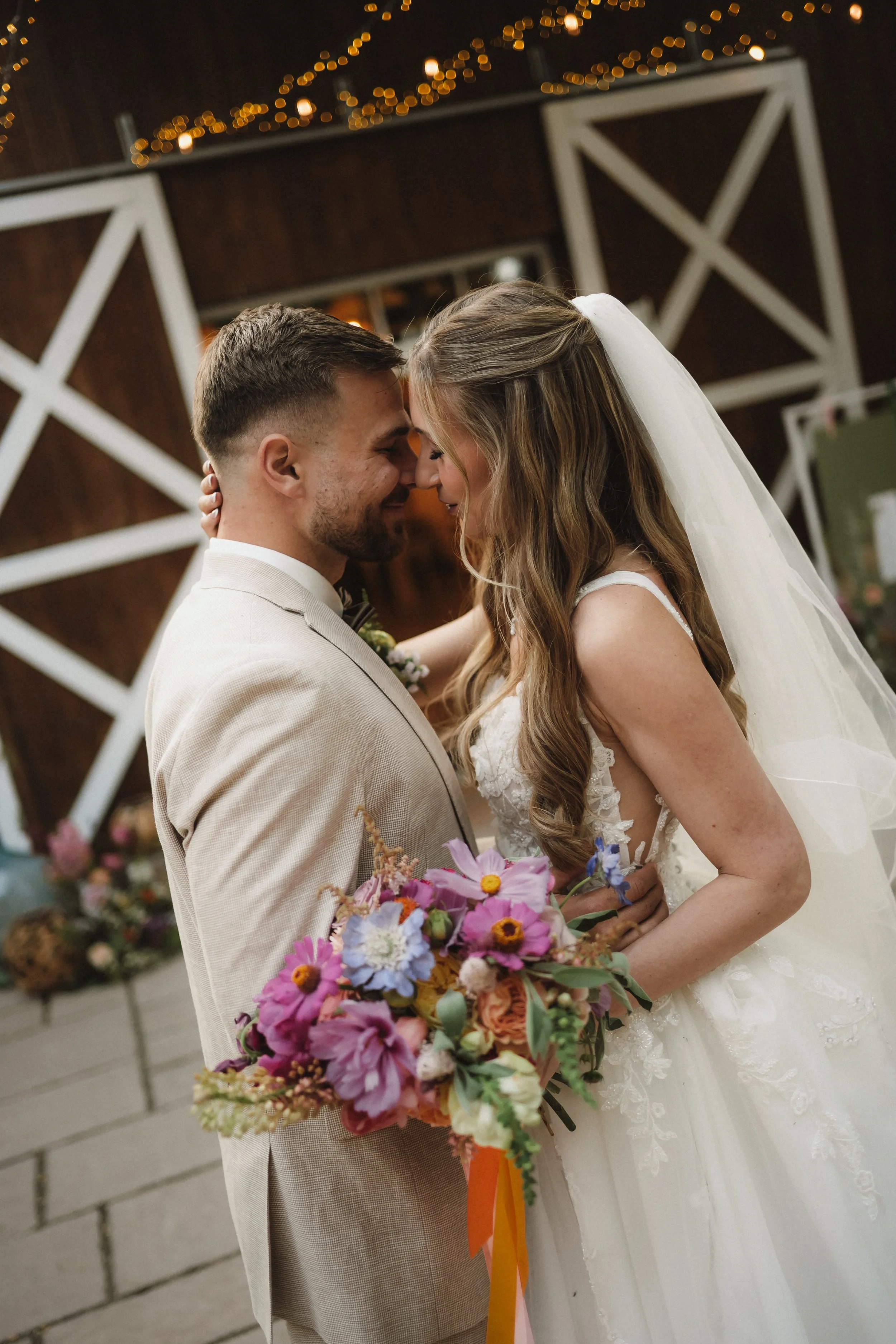 A menyasszony és a vőlegény meghitt pillanatot osztanak meg az esküvőjükön, homlokukat egymáshoz érintve, mosolyogva. A menyasszony fehér csipkeruhát és fátylat visel, kezében színes virágcsokorral, a vőlegény világos öltönyt hord. 