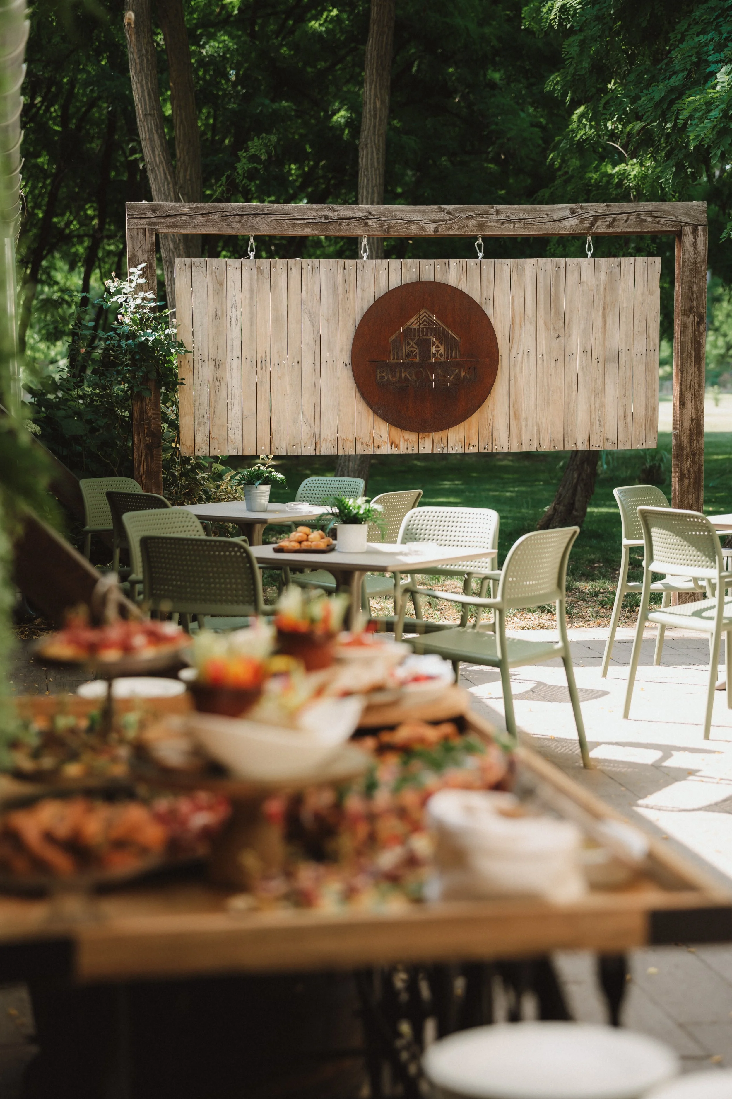 Outdoor dining area with tables and chairs, a buffet table with food, and a wooden sign with a barn and the word "BUKOWSKI" on a large wooden frame surrounded by trees.