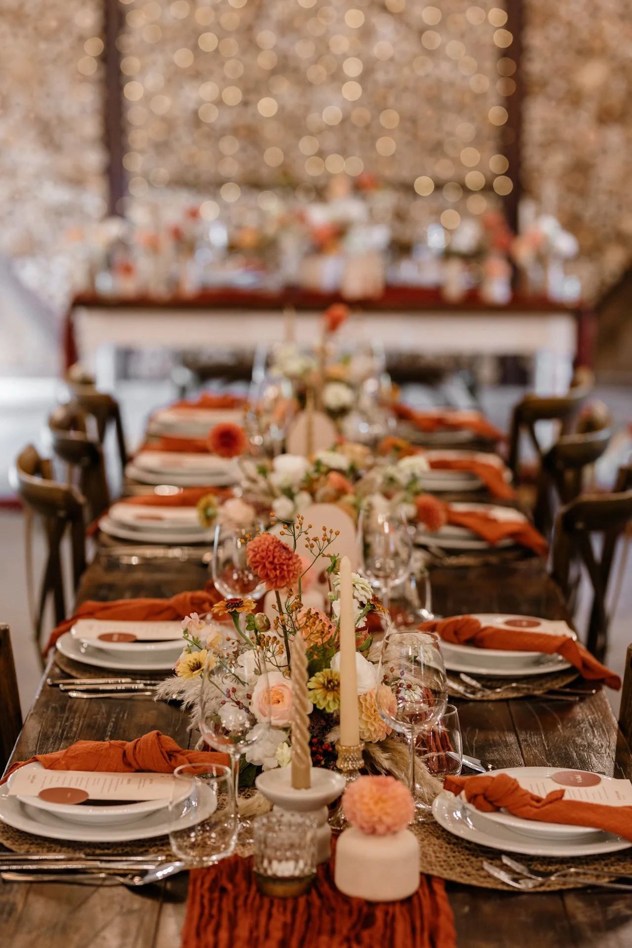 Egy hosszú, rusztikus fa esküvői asztal meleg és romantikus hangulatot teremt. A dekoráció narancs és barack árnyalatú virágokból, gyertyákból és elegáns terítékből áll, amelyek harmonikusan egészítik ki a természetes fa textúráját. 
