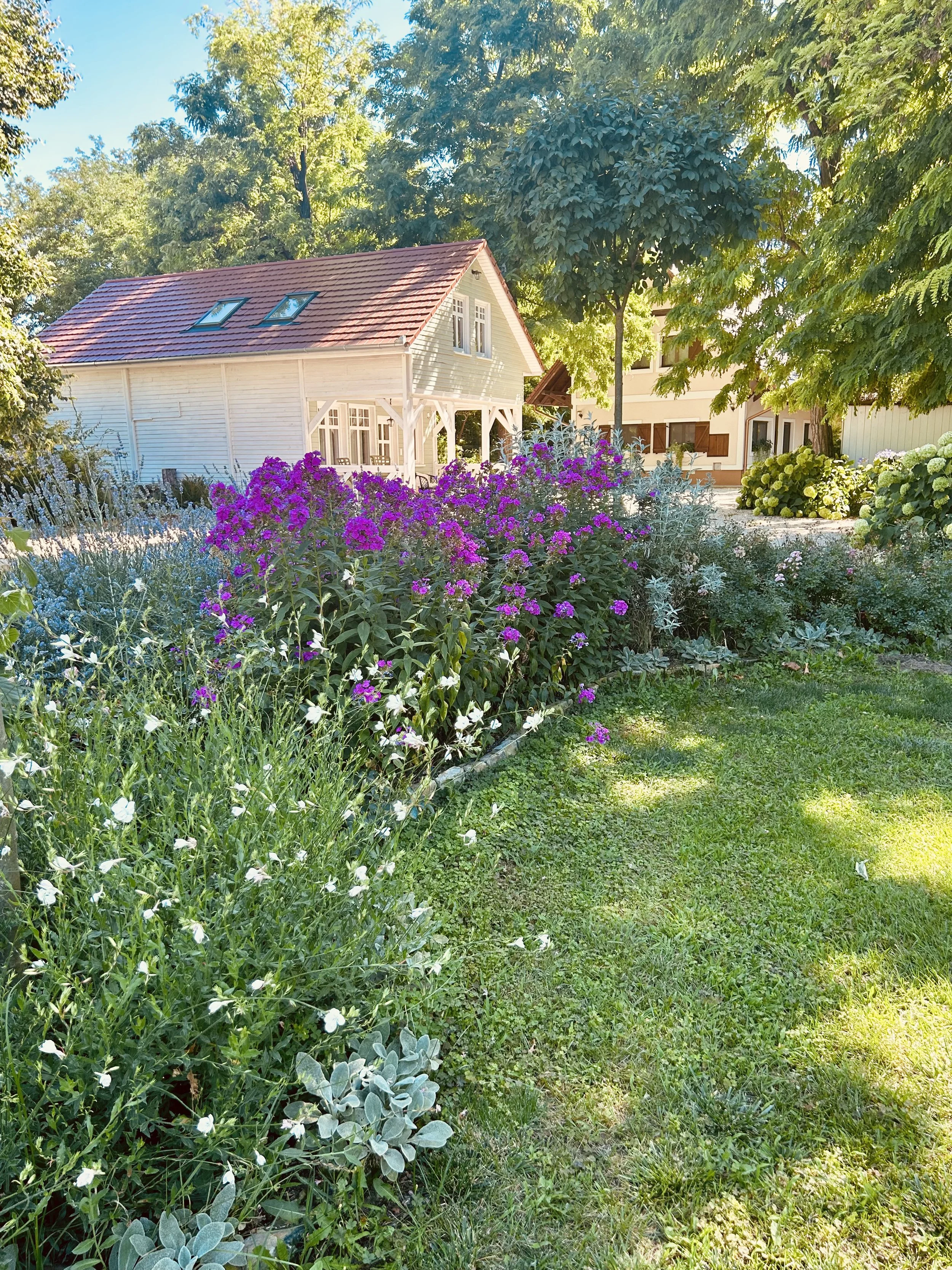 A garden with colorful flowers, green grass, and trees, with a white house with a red roof in the background.