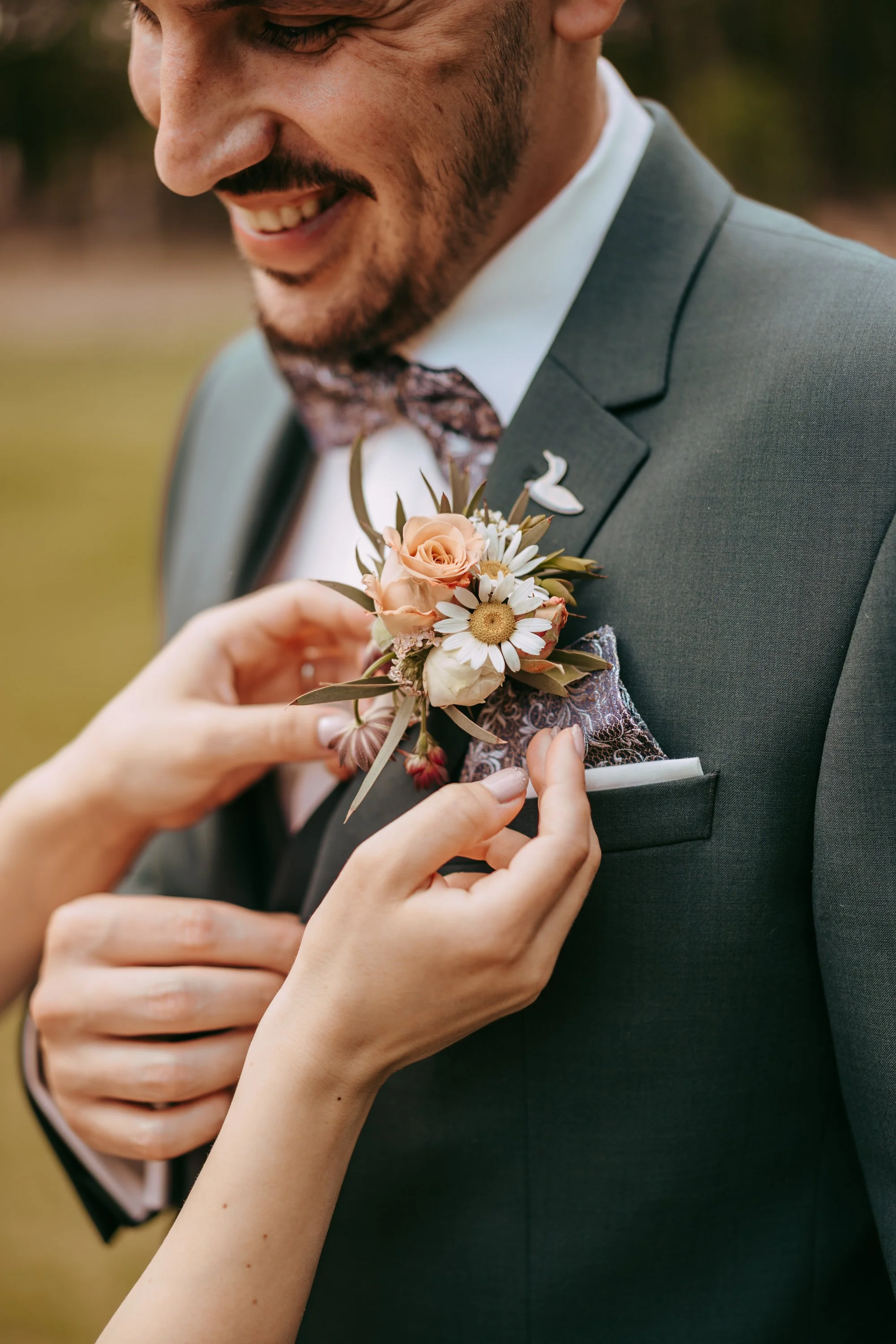 A person in a gray suit is being helped to pin a boutonniere with peach roses, white daisies, and green leaves on their lapel.