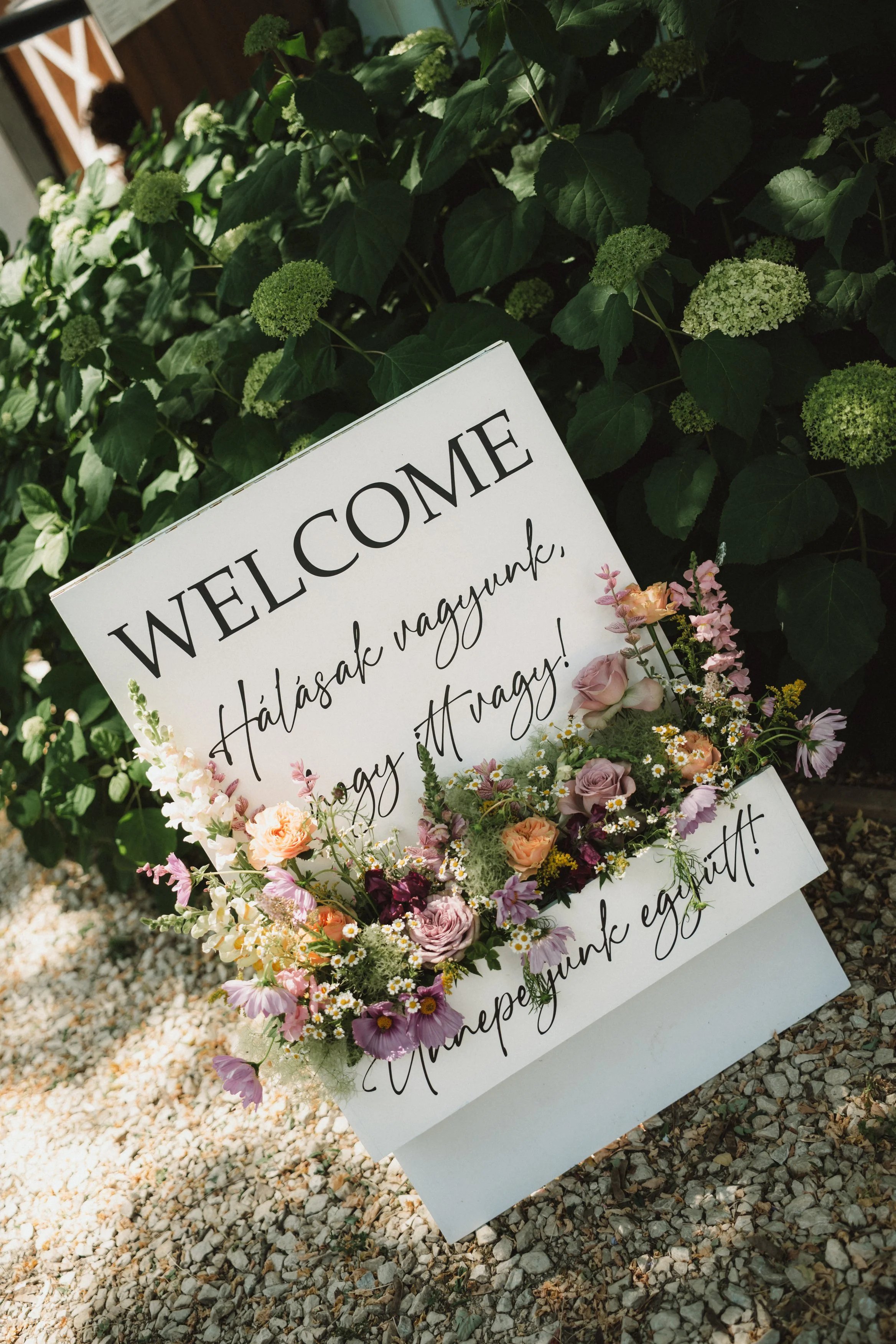 Decorative welcome sign with a floral border, placed on gravel ground in front of green bushes.