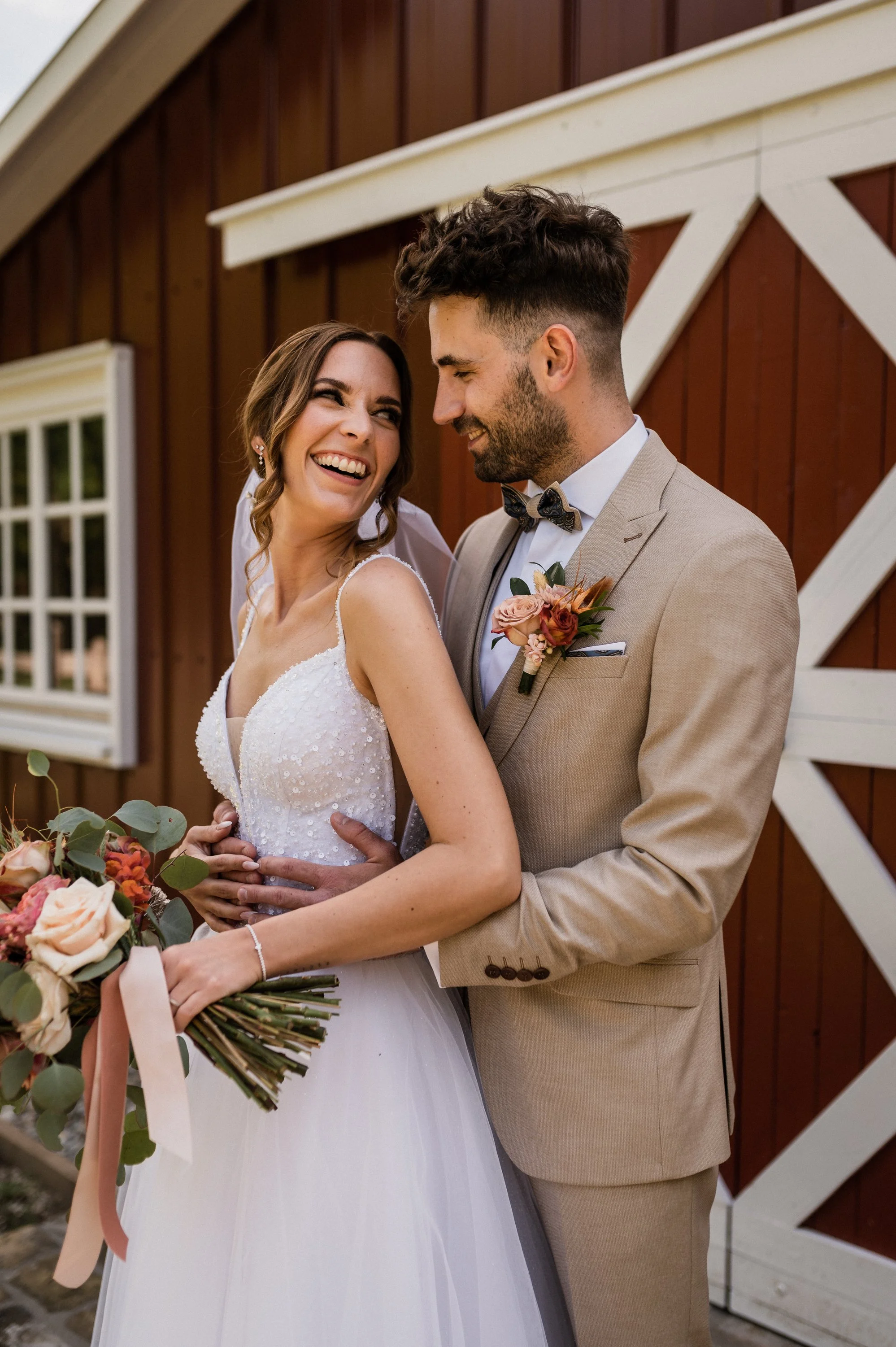 Egy boldog menyasszony és vőlegény ölelkezve néznek egymásra, miközben egy vörös rusztikus fa pajta előtt állnak.
