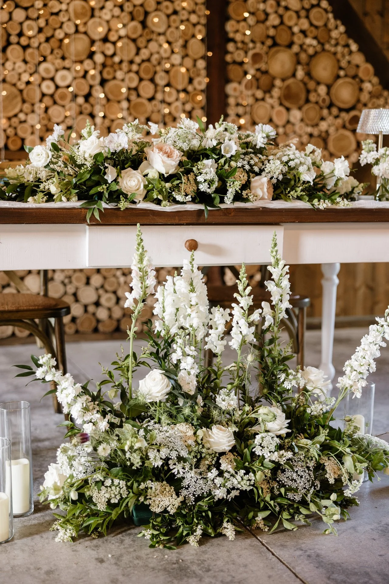 Fehér virágkompozíció asztalon, gyertyákkal, rusztikus fa fal háttérrel.
