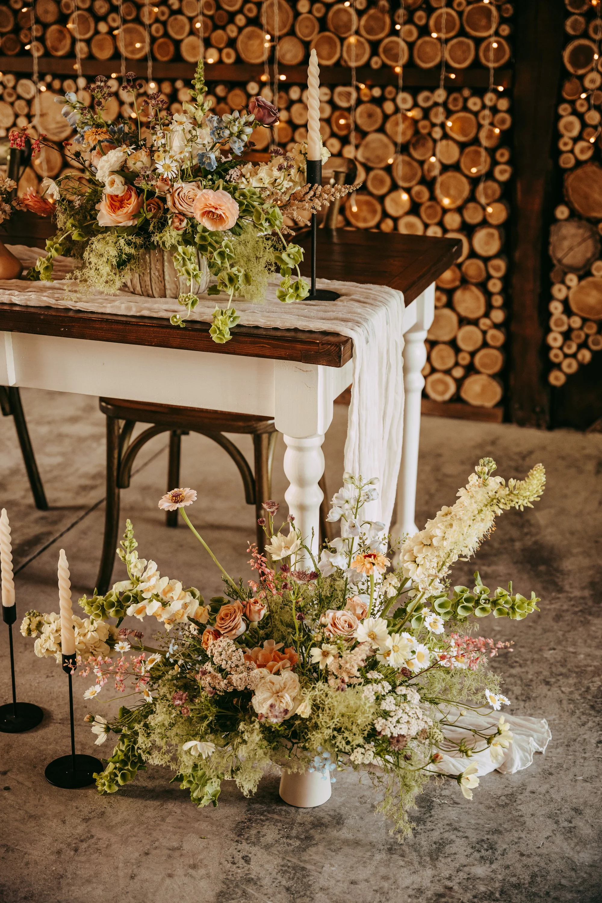 Decorated wedding or event table with floral arrangement and candles, set against a background of stacked firewood.