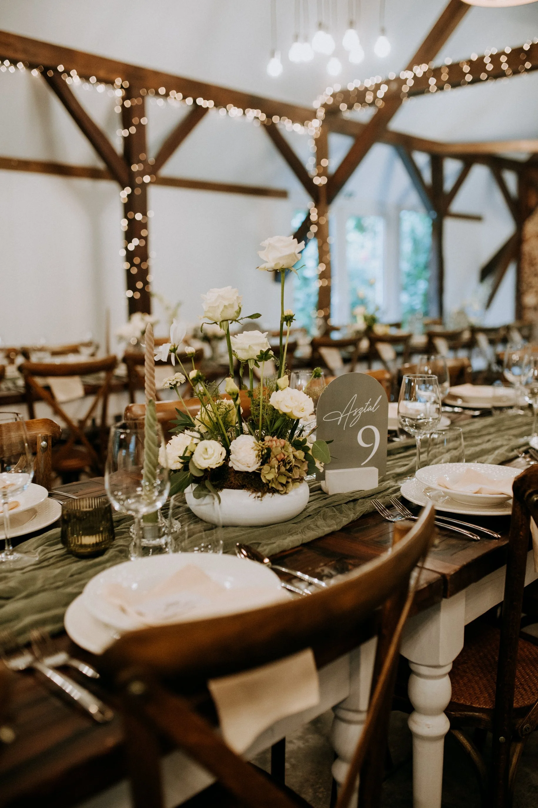 A decorated wedding reception table with a floral centerpiece, table number '9', and glassware, set in a rustic room with wooden beams and string lights.
