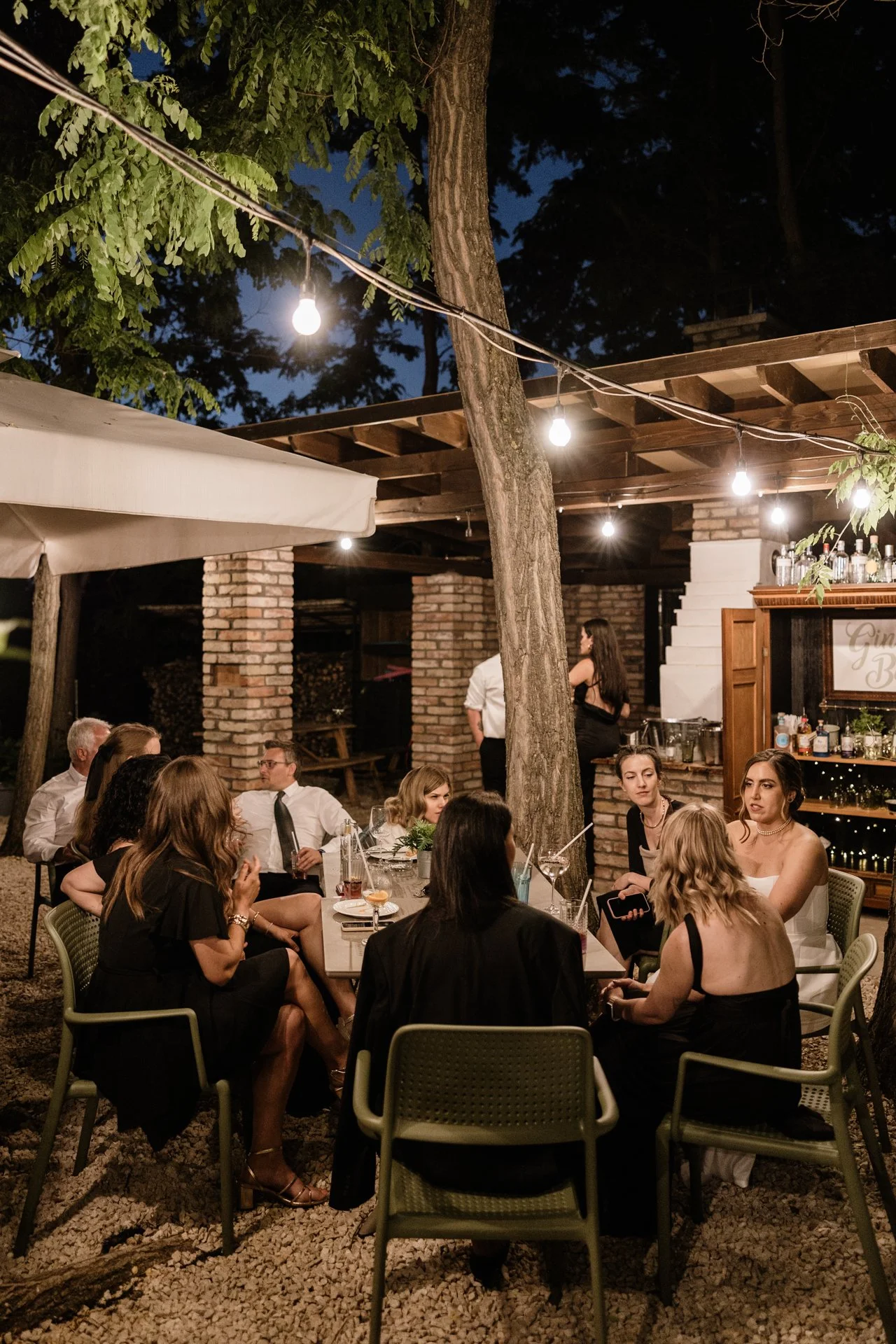 People gathered around a table at an outdoor nighttime party under string lights, with trees, brick pillars, and a bar in the background.