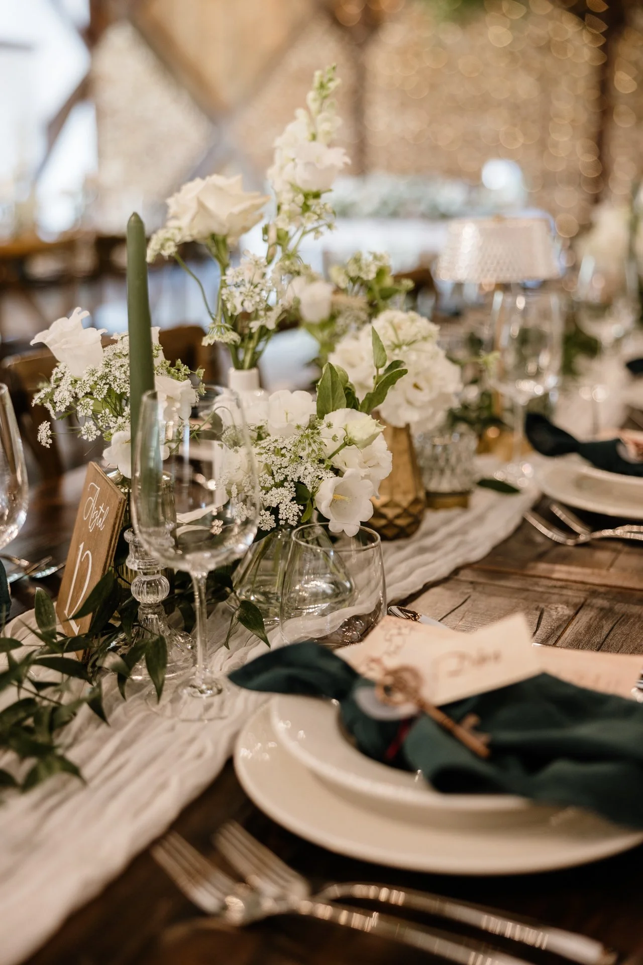 Esküvői fogadásra terített asztal fehér virágokkal, gyertyákkal, poharakkal és terítékkel, köztük egy szalvétával, rajta gyűrű és egy kis kártya.
