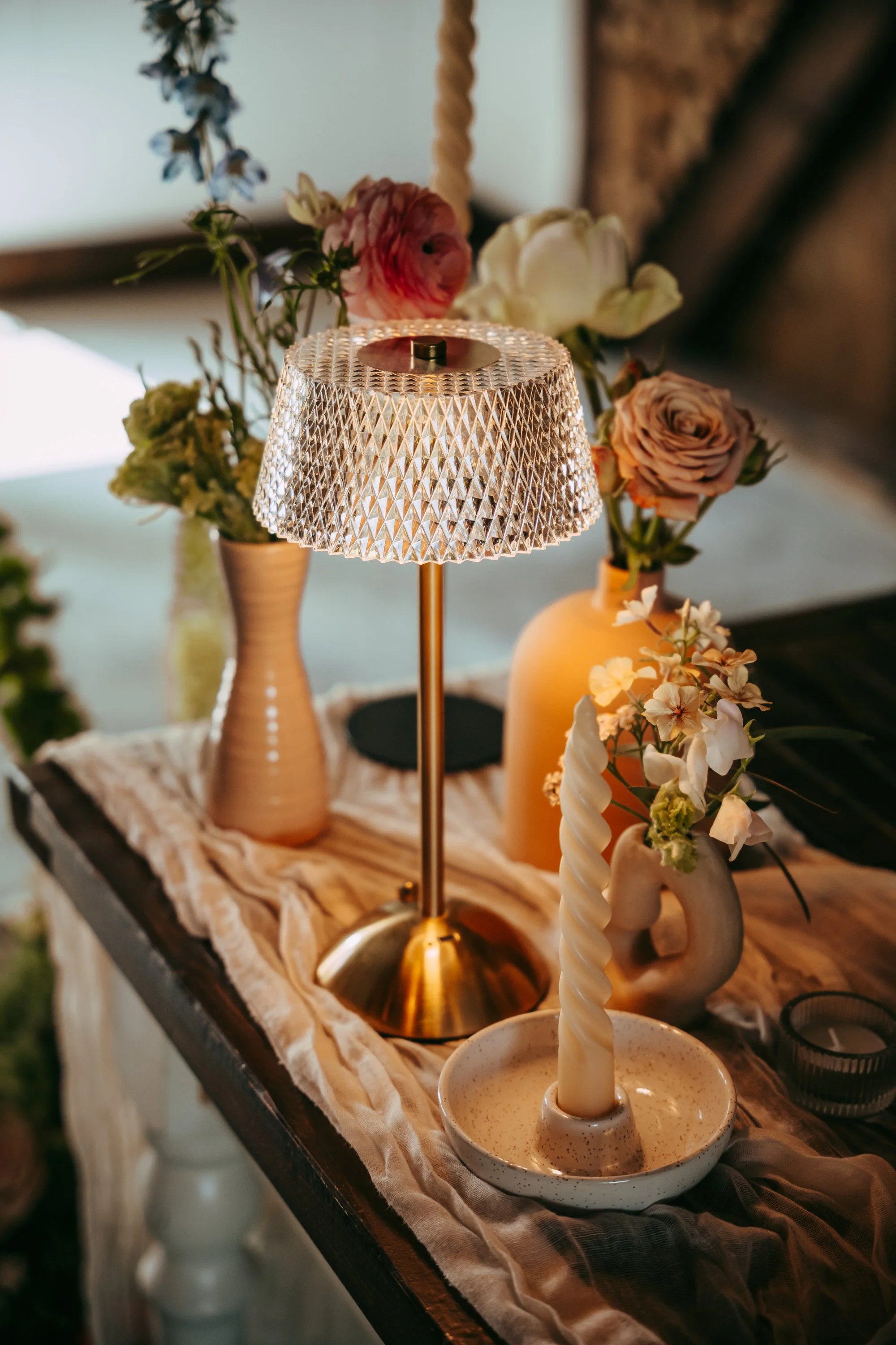 A table decorated with various vases and a lamp, with a cream-colored cloth, and filled with flowers including roses and other blooms.