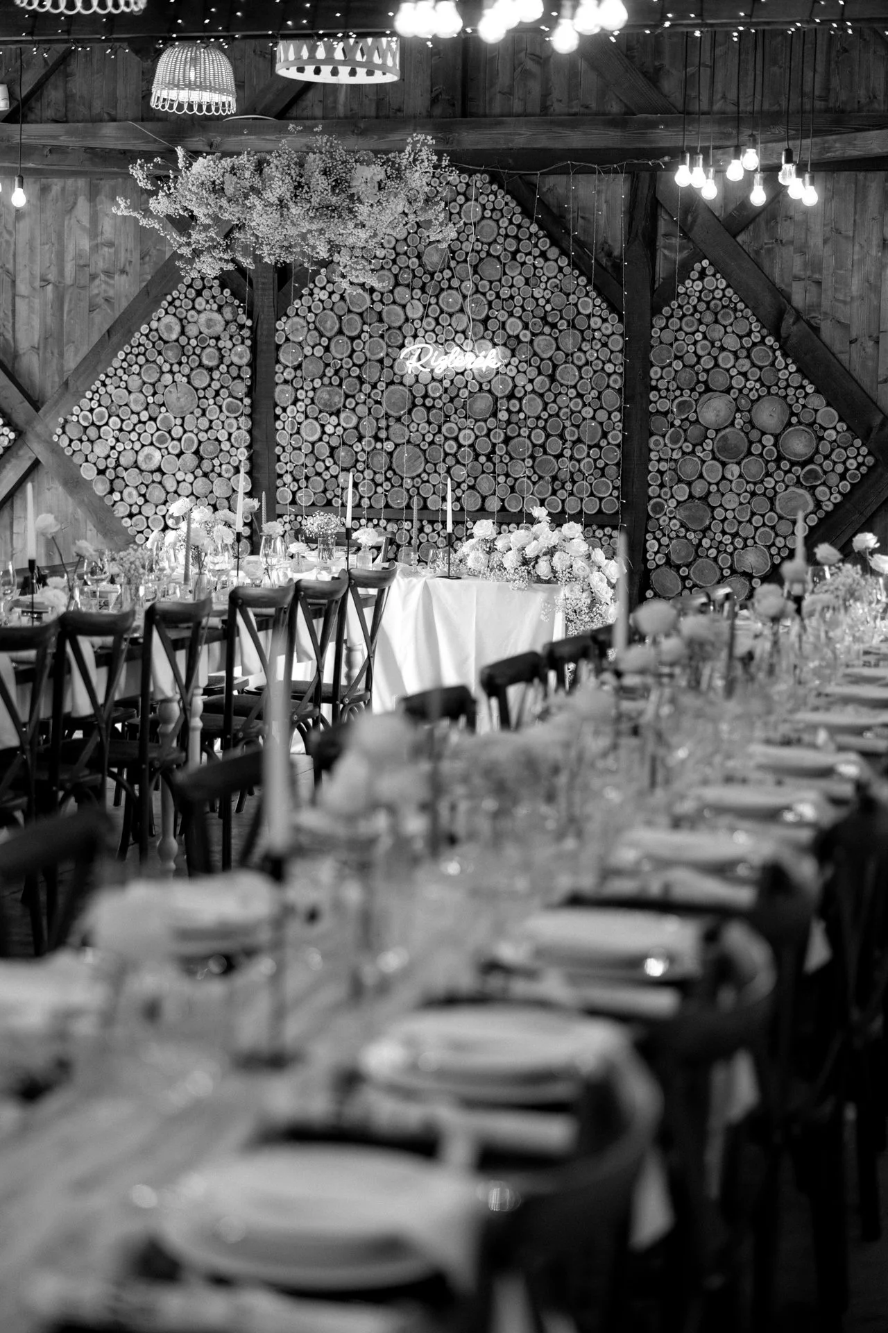 Fekete-fehér hangulatú fotó egy elegáns esküvői vacsoráról. A hosszú asztal elegánsan megterítve áll tányérokkal, poharakkal és virágos asztaldíszekkel. A háttérben egy dekorált rönkfa fal látható, amelyen egy világító neon felirat