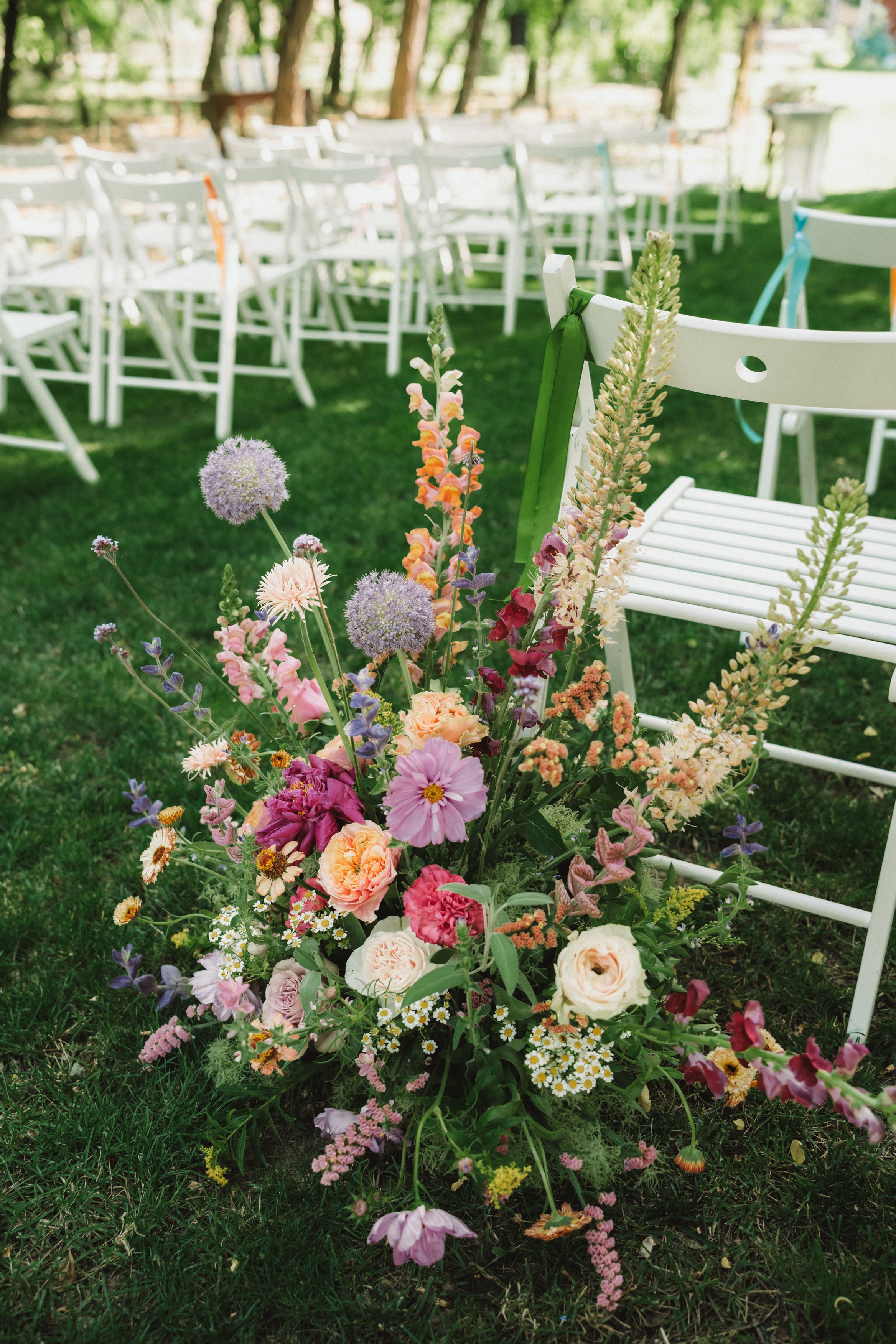 Colorful flower arrangement on green grass near white chairs decorated with ribbons at an outdoor wedding venue.