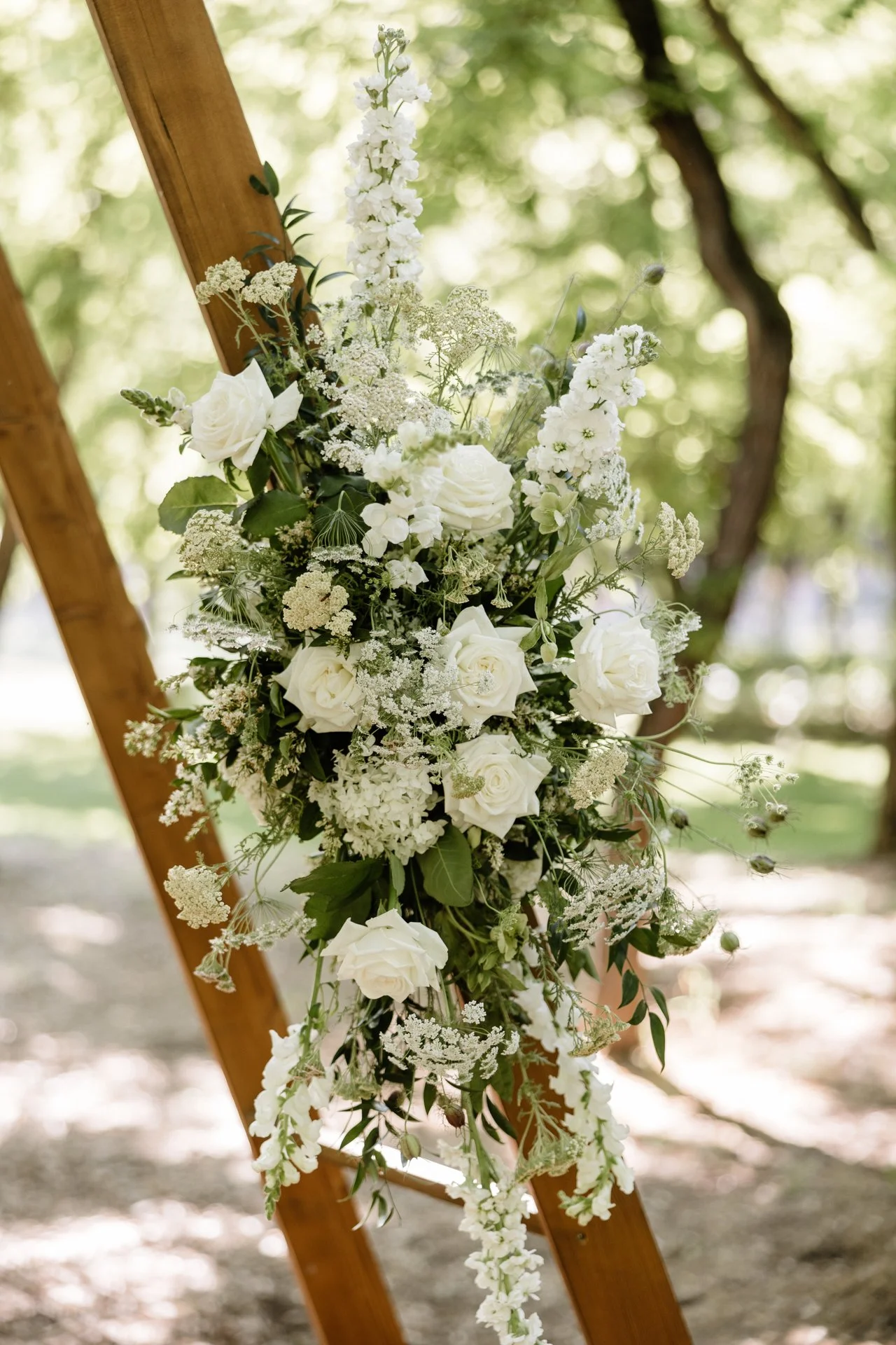 Fehér virágkompozíció rózsákkal, szarkalábbal és más virágokkal, fa szerkezetre rögzítve szabadtéren, háttérben zöld fákkal.
