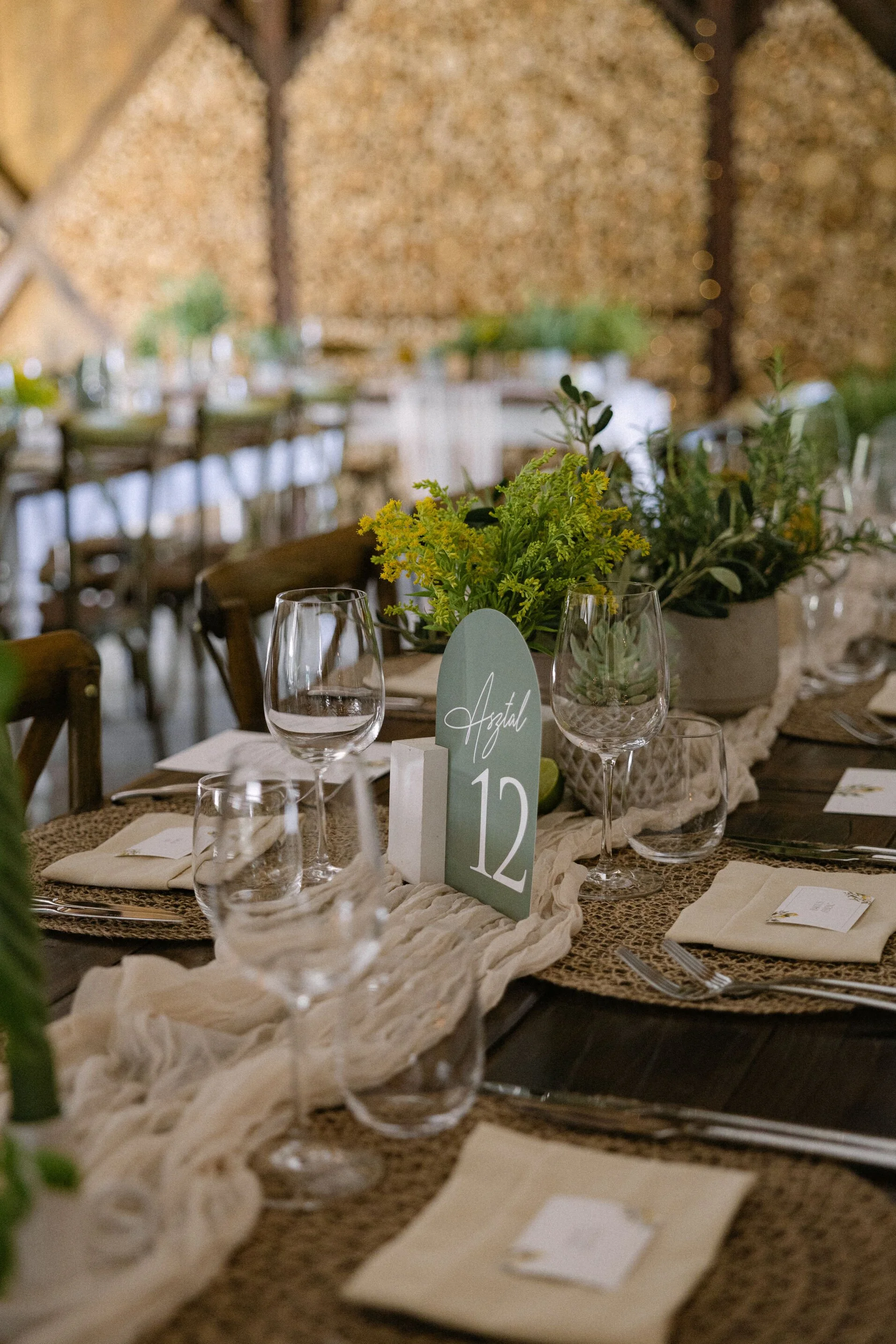 Elegant dining table setup with glassware, napkins, and table number card with the name 'Azal' and the number '12'. Decorated with green plants and a cream-colored fabric runner.