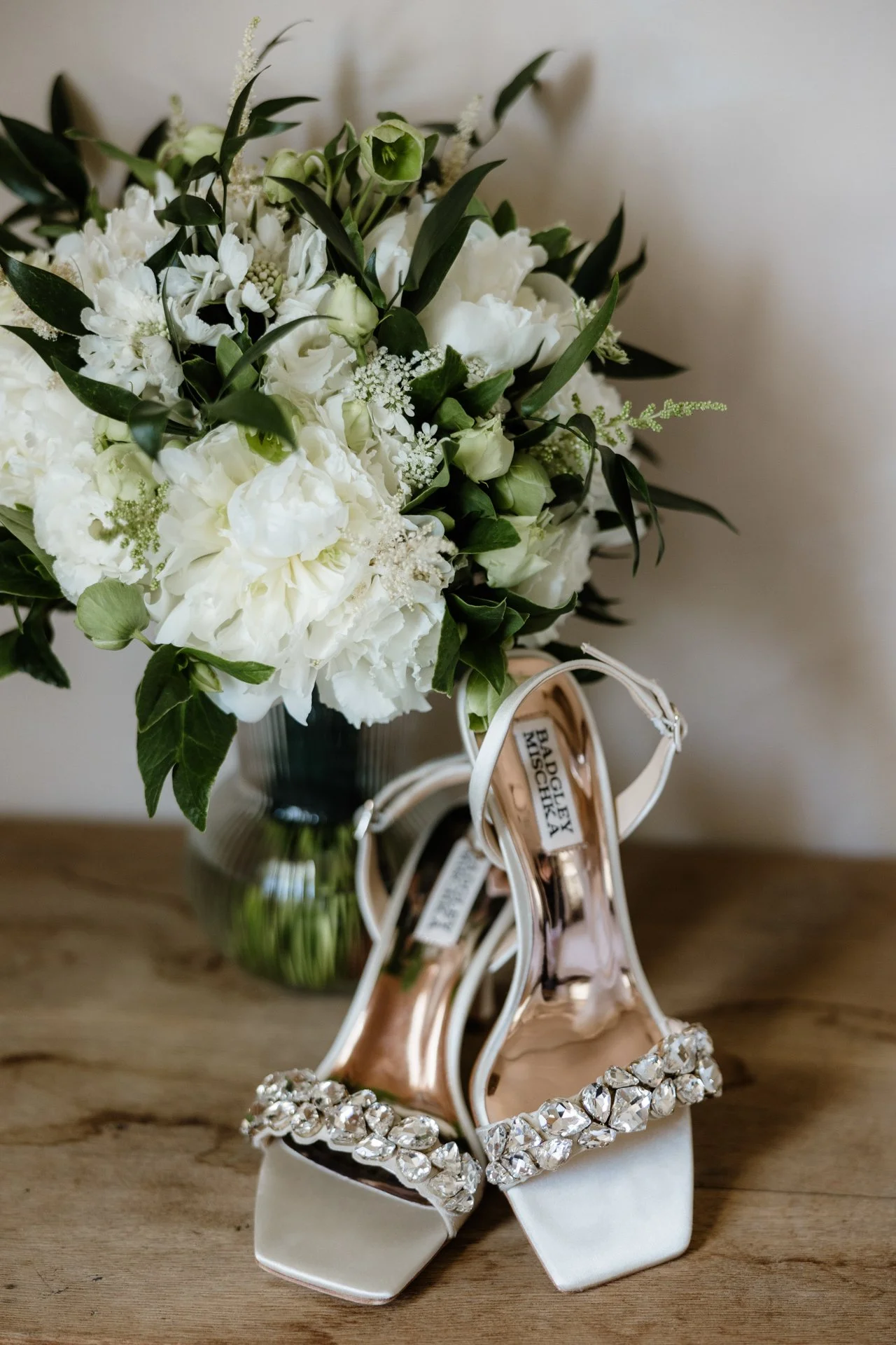 A bouquet of white flowers in a vase and a pair of white high-heeled shoes with rhinestone embellishments on a wooden surface.