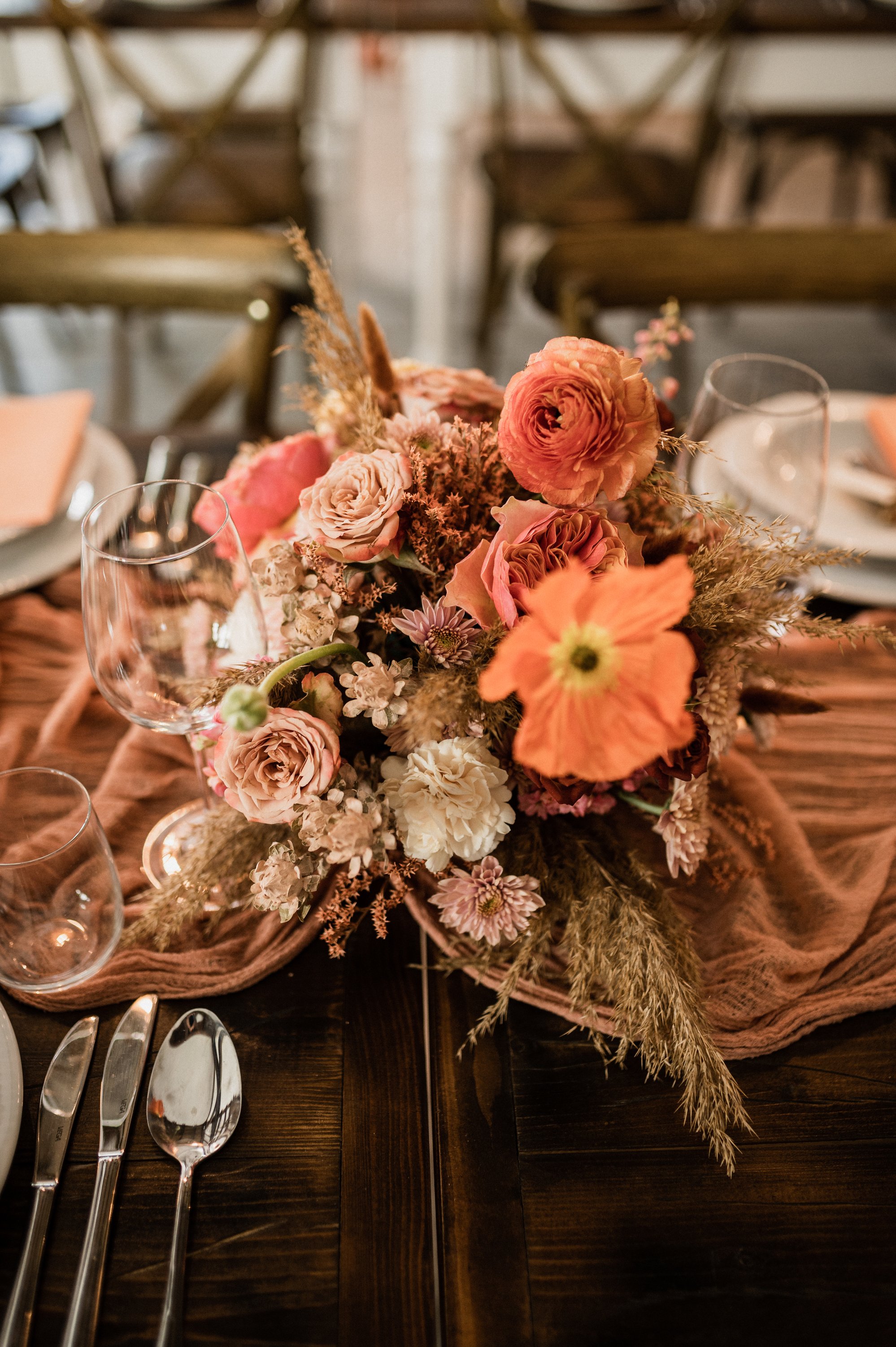 A floral centerpiece with pink, peach, and cream flowers on a dining table with glasses, silverware, and pink cloth napkins.