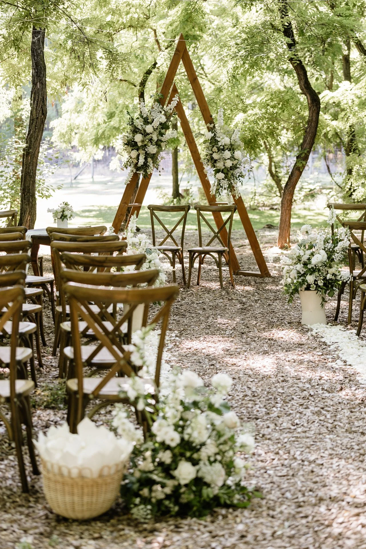 Szabadtéri esküvői helyszín fa székekkel, virágkompozíciókkal és egy háromszög alakú fa ívvel, amelyet fehér virágokkal díszítettek, erdős környezetben.
