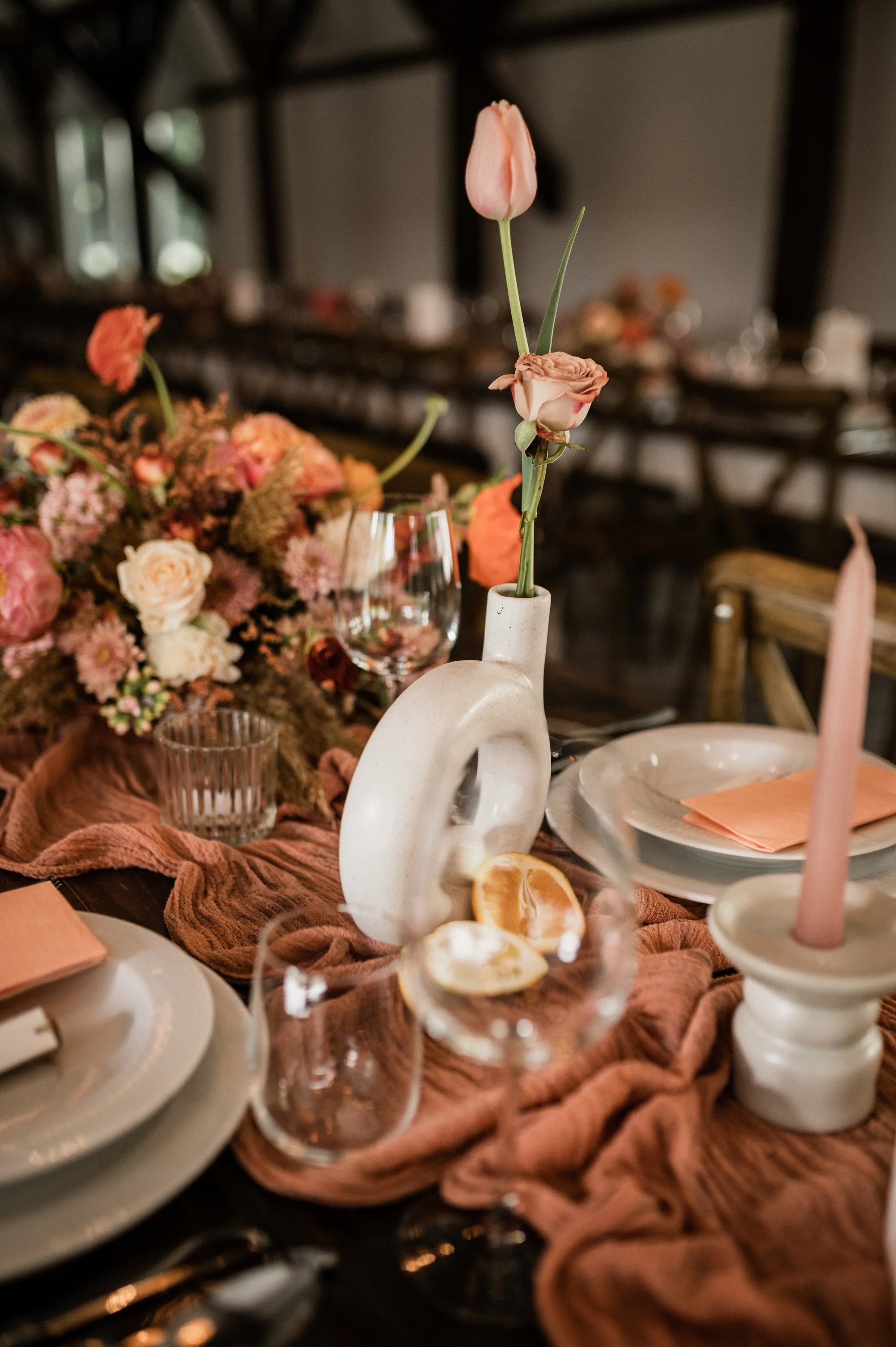 Table setting with a white vase holding pink and peach roses and tulips, surrounded by pink and orange flowers, candles, and glassware on a table with a mauve tablecloth.