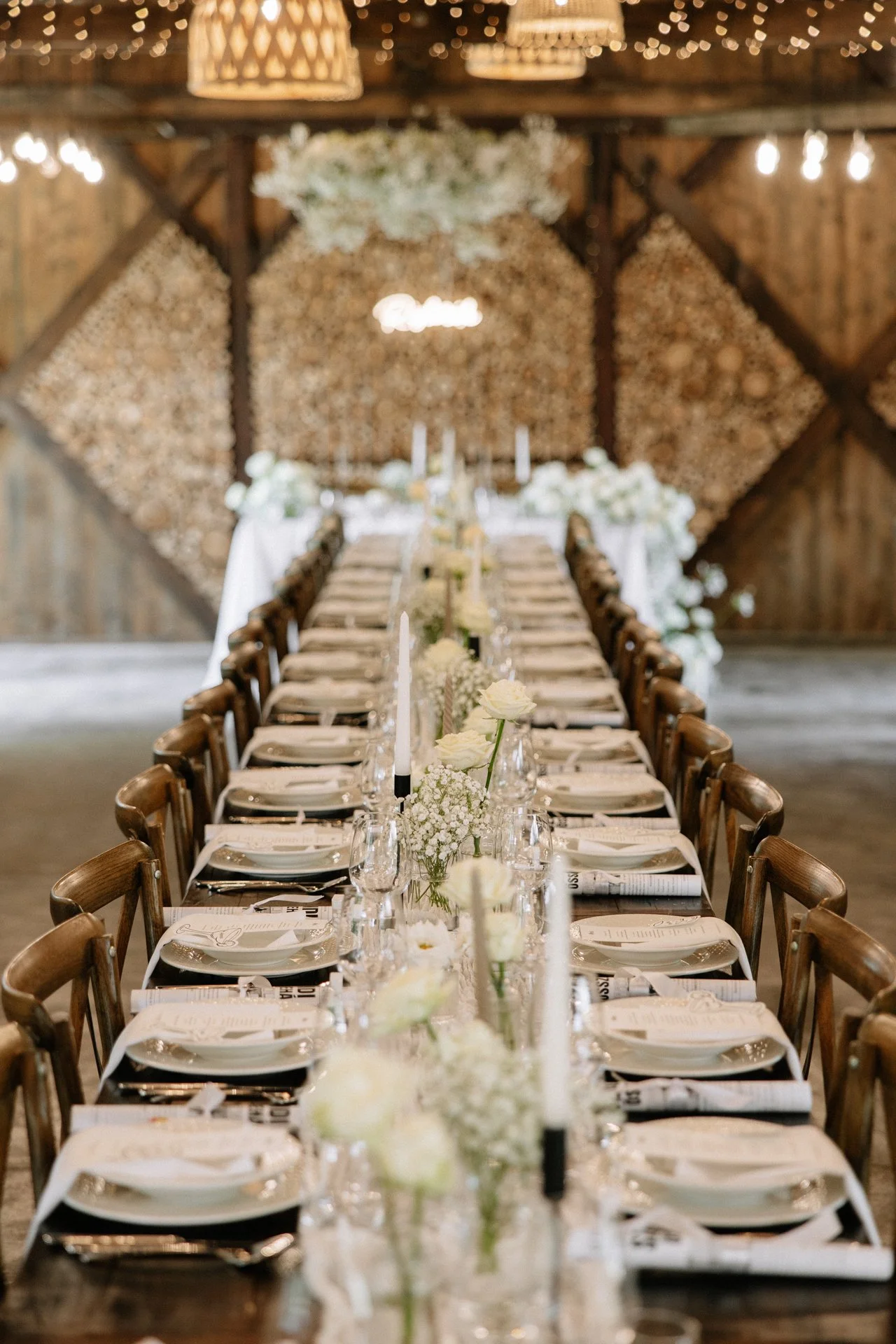 Hosszú, téglalap alakú étkezőasztal ünnepi alkalomra megterítve fehér virágokkal, gyertyákkal és elegáns terítékkel, egy elegáns pajta környezetben, fa falakkal és mennyezetről lelógó fényfüzérekkel.
