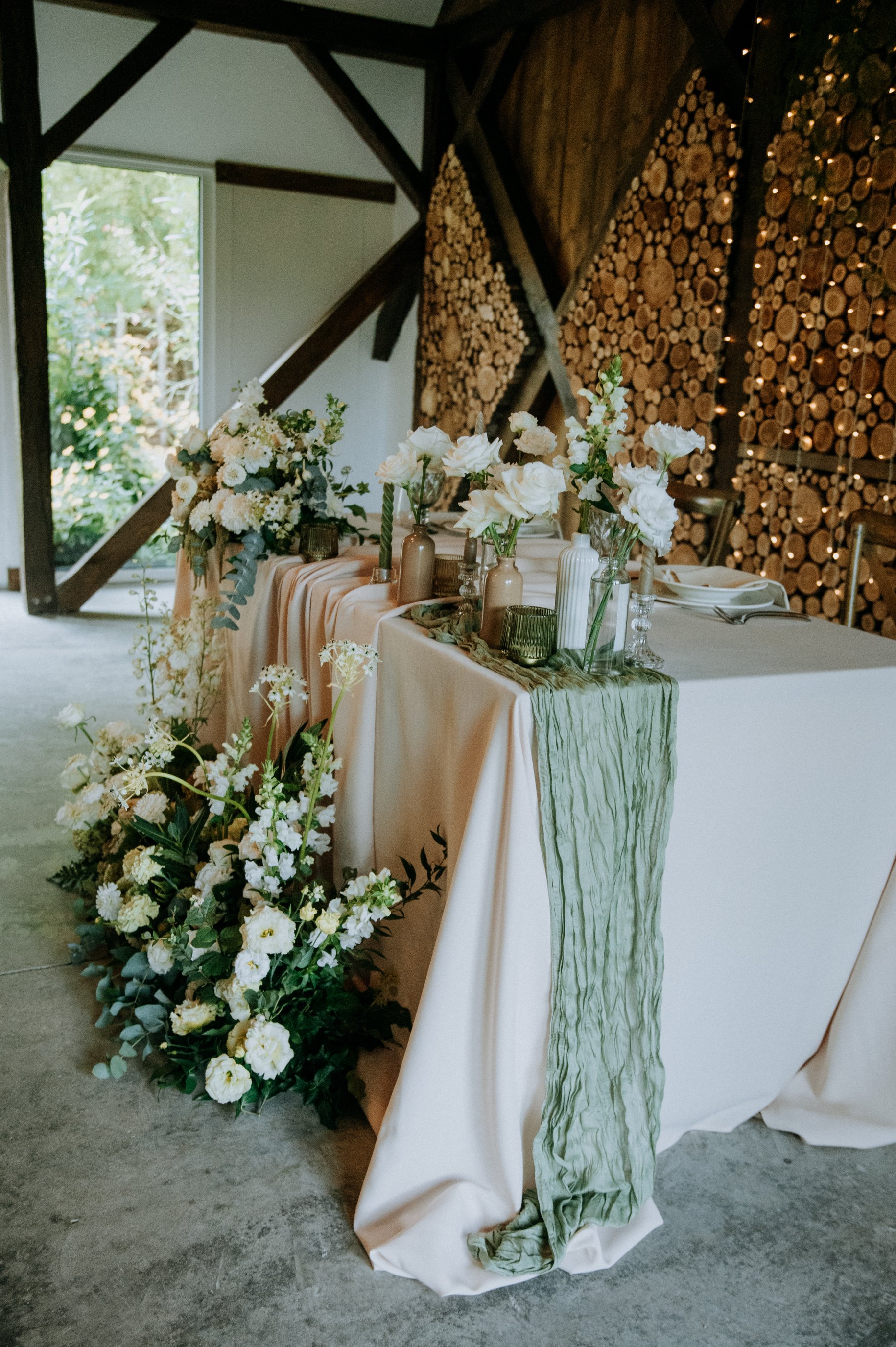 Esküvő  asztal fehér és bézs virágokkal, zöld növényzettel és vázákkal, zöld asztali futóval, háttérben rönkfa fal és pajta belső tér.
